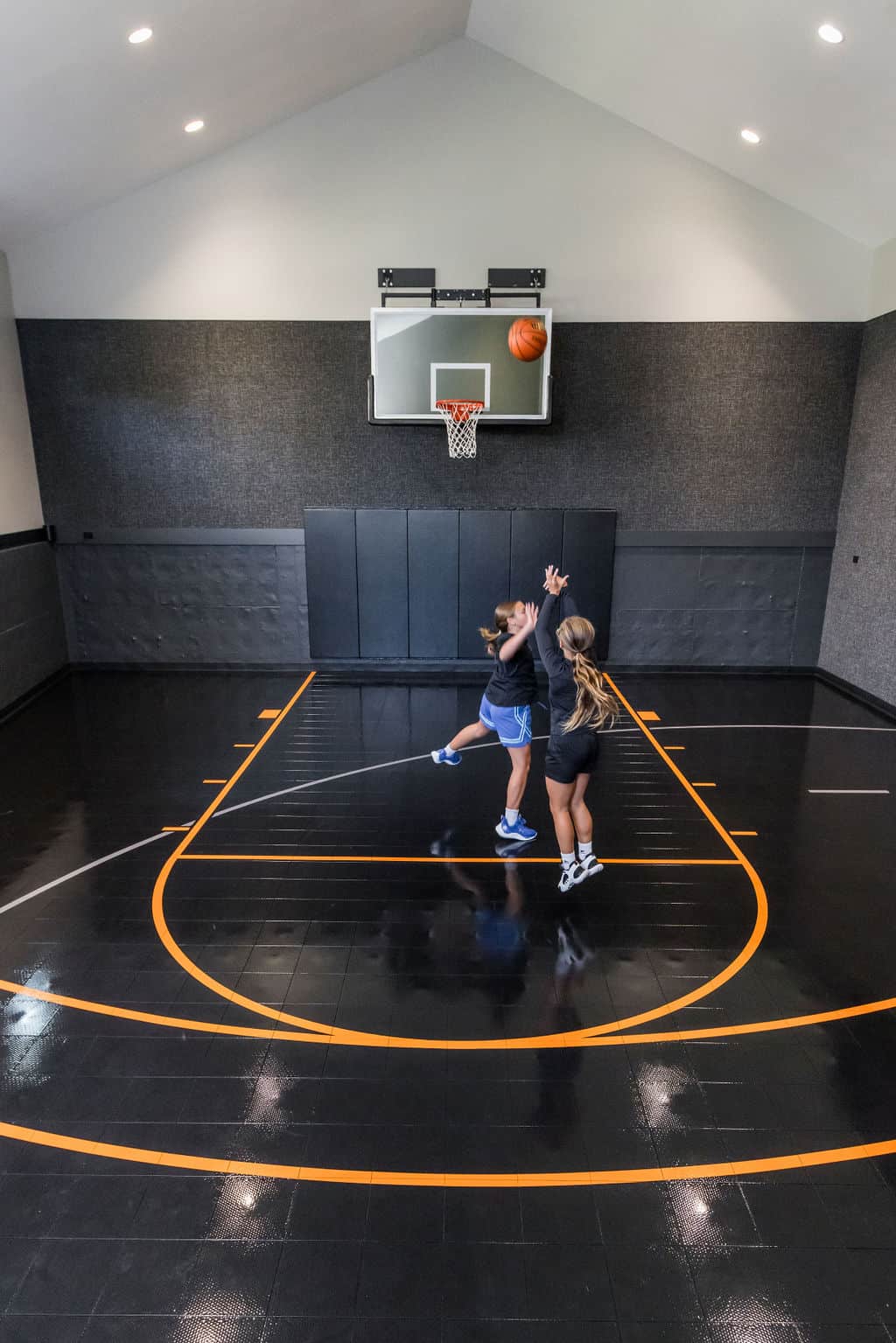 Nicholas Design Build | Two people play basketball on an indoor court. One person is jumping to shoot the ball towards the hoop, while the other attempts to block.