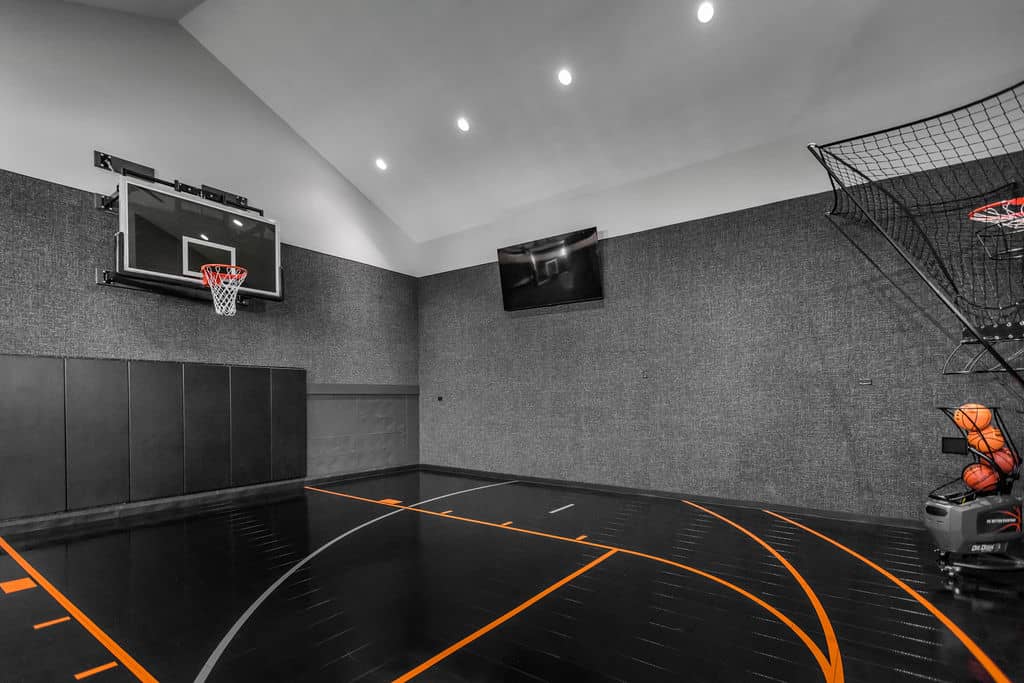 Nicholas Design Build | Indoor basketball court with a hoop, black flooring, and a ball return machine. Two screens are mounted on the wall.