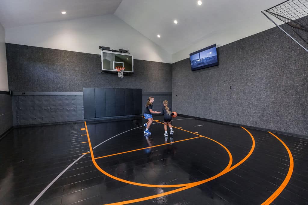 Nicholas Design Build | Two people playing basketball in an indoor court with dark flooring and gray walls. A hoop and a wall-mounted screen are visible.