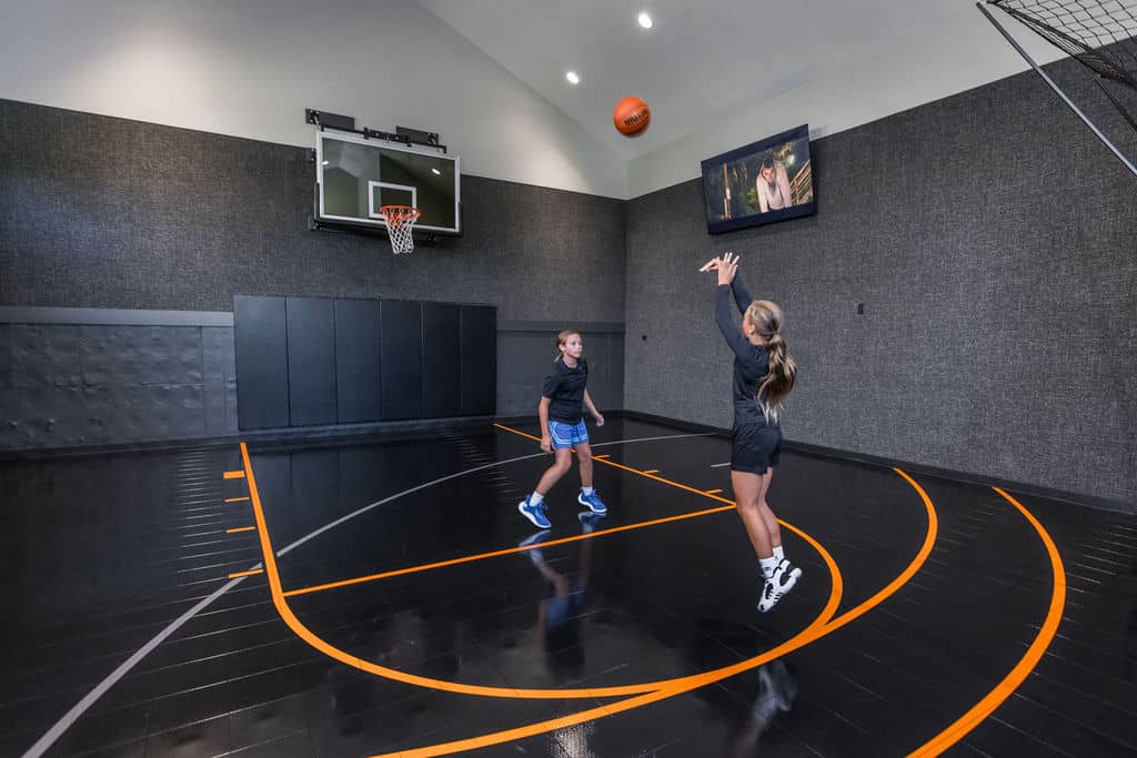 Nicholas Design Build | Two people playing basketball indoors. One person is shooting the ball while the other watches. There's a TV and a basketball hoop on the wall. The court has black flooring with orange lines.