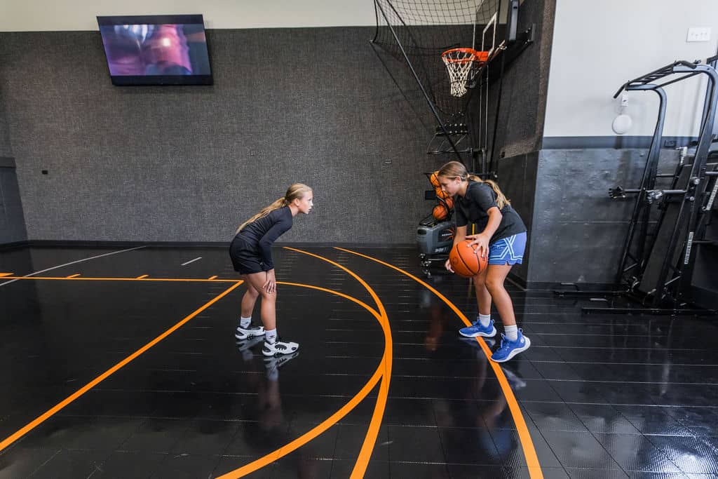 Nicholas Design Build | Three people playing basketball indoors on a black court. Two are preparing to pass or shoot the ball while one is defending. A TV is on the wall in the background.