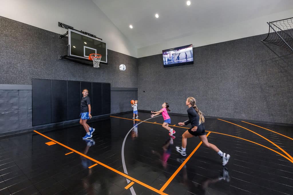 Nicholas Design Build | Four people are playing basketball indoors on a black court. Two adults and two children are in motion near the hoop.