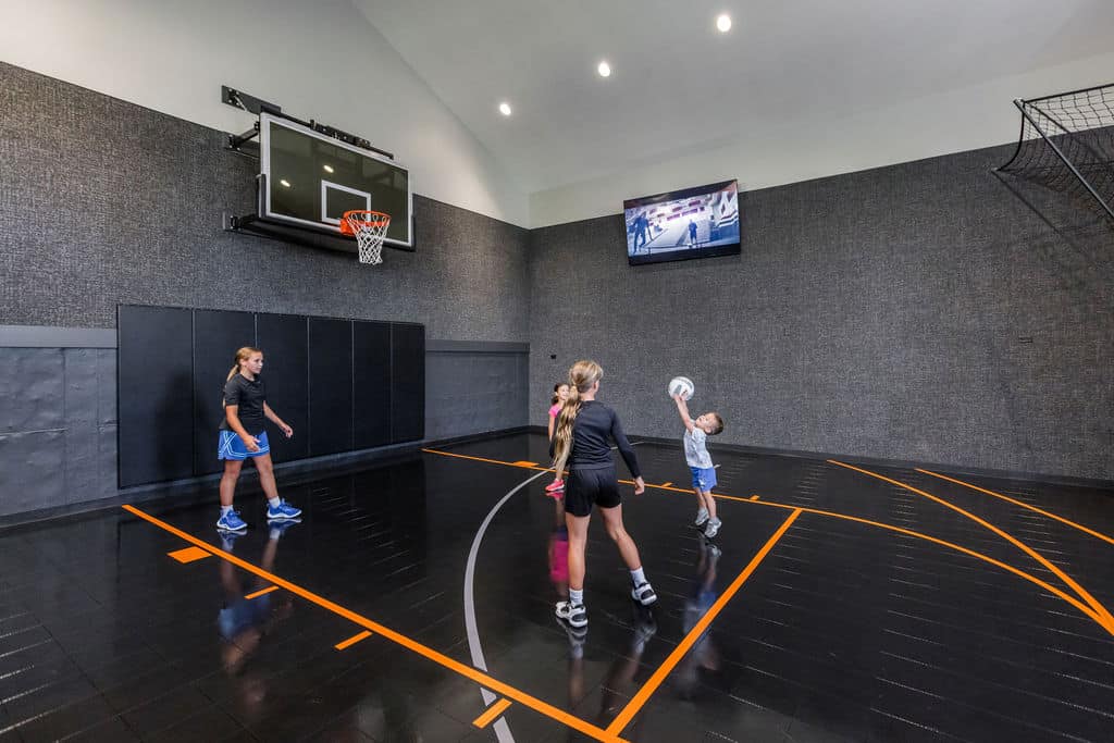 Nicholas Design Build | Three children play basketball indoors on a shiny black court with orange lines. A scoreboard and screen are visible on the dark gray walls.