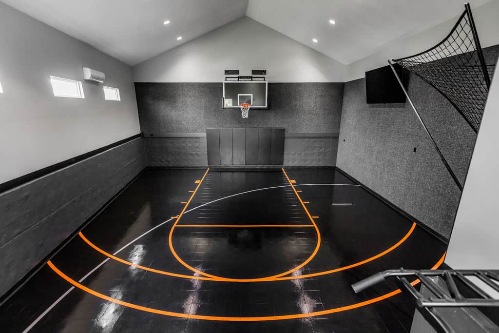 Nicholas Design Build | Indoor basketball court with a hoop, high ceiling, gray walls, and black floor featuring orange lines. A basketball return net is attached to the hoop.