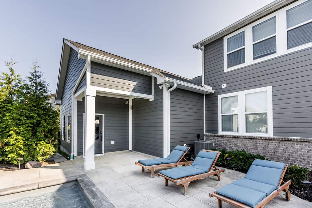 Nicholas Design Build | Backyard view of a modern gray house with a small pool, patio area, and three wood-framed blue lounge chairs.