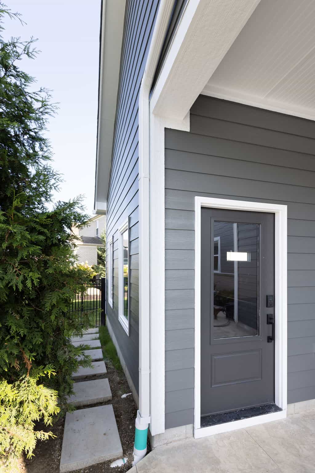 Nicholas Design Build | Side view of a modern gray house with a narrow pathway and a dark gray door, adjacent to a row of green shrubs.