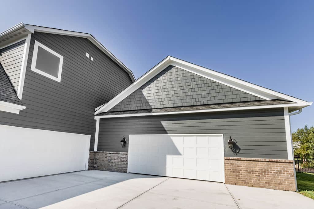 Nicholas Design Build | Garage doors on a modern gray and white house, featuring brick accents and two outdoor wall lights, under a clear blue sky.