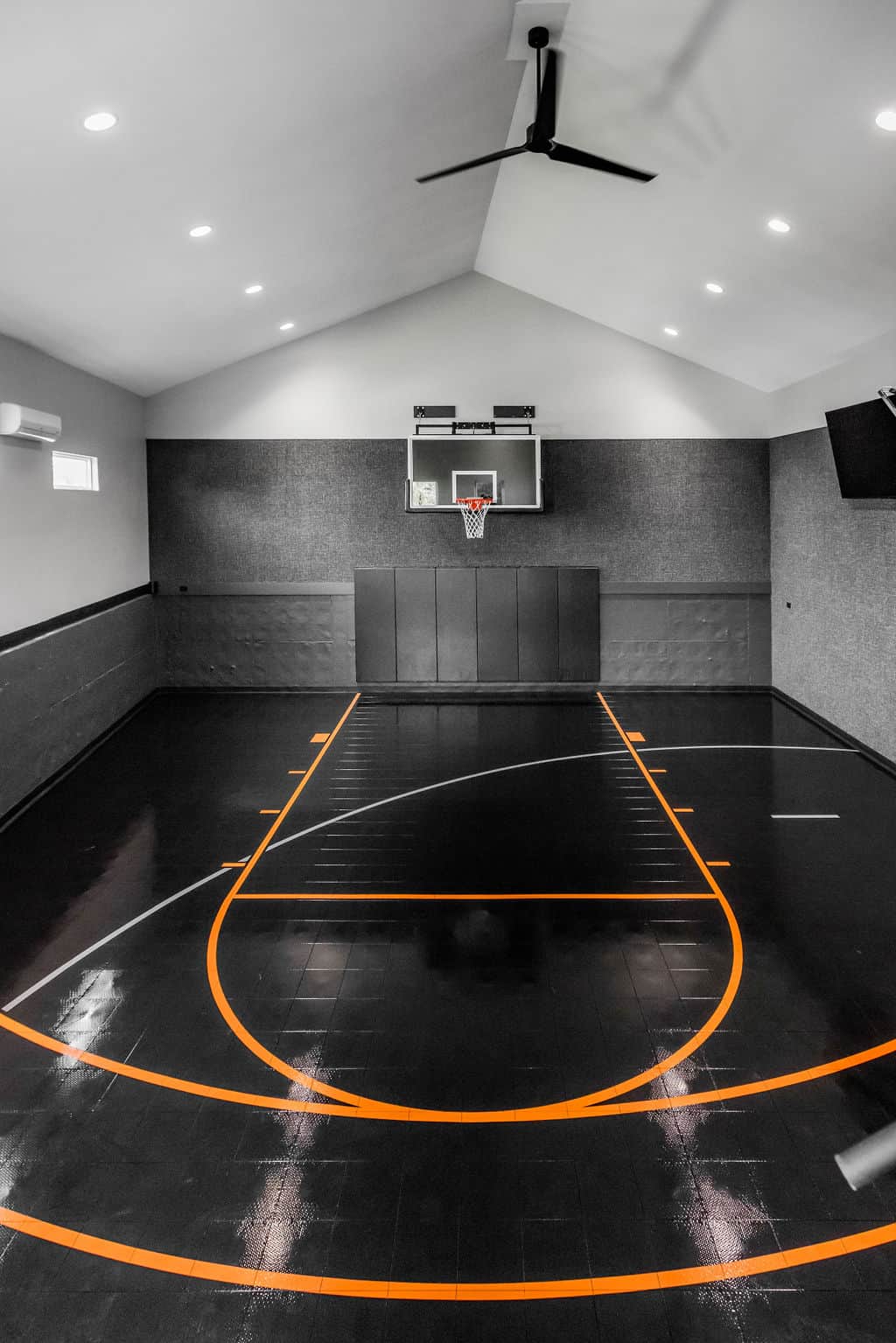 Nicholas Design Build | Indoor basketball court with black flooring and orange lines, featuring a single hoop and a ceiling fan.