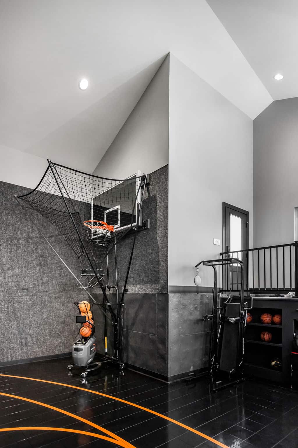 Nicholas Design Build | Indoor basketball hoop with a ball return net system, a ball rack, and a black and orange court floor.