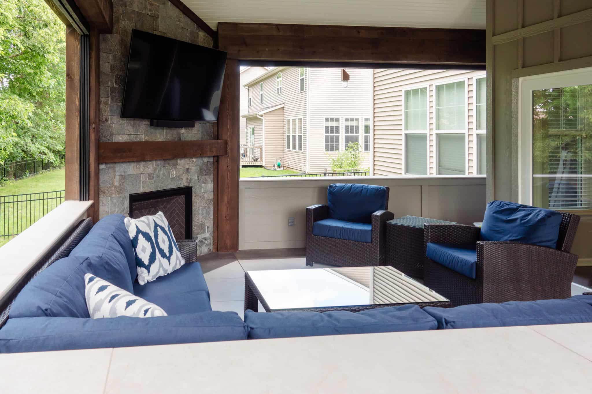 Nicholas Design Build | A covered outdoor patio features a seating area with blue cushions, a TV mounted above a stone fireplace, and large windows overlooking a grassy yard.