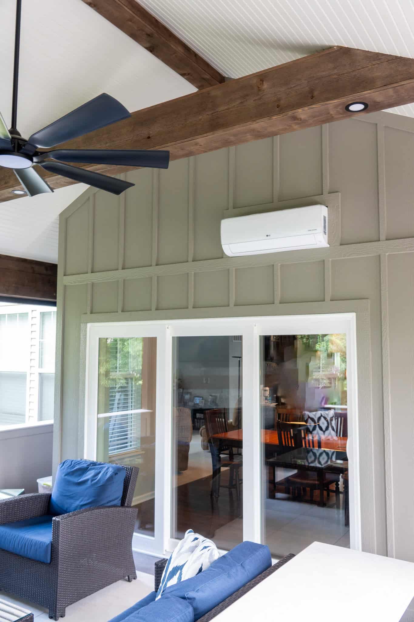 Nicholas Design Build | Room with a ceiling fan, wooden beams, and air conditioning unit. Glass doors open to reveal a dining area with a table and chairs. Blue and brown furniture visible on the patio.