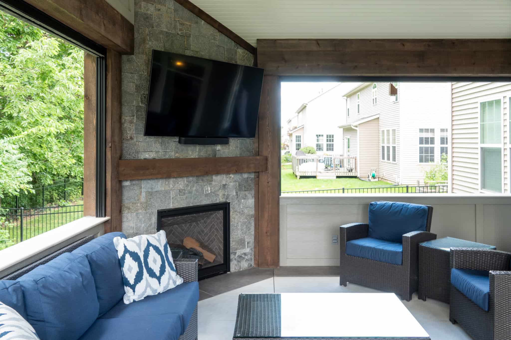 Nicholas Design Build | A patio with blue cushioned seating, a stone fireplace, and a mounted TV. Large windows offer a view of neighboring houses and greenery.