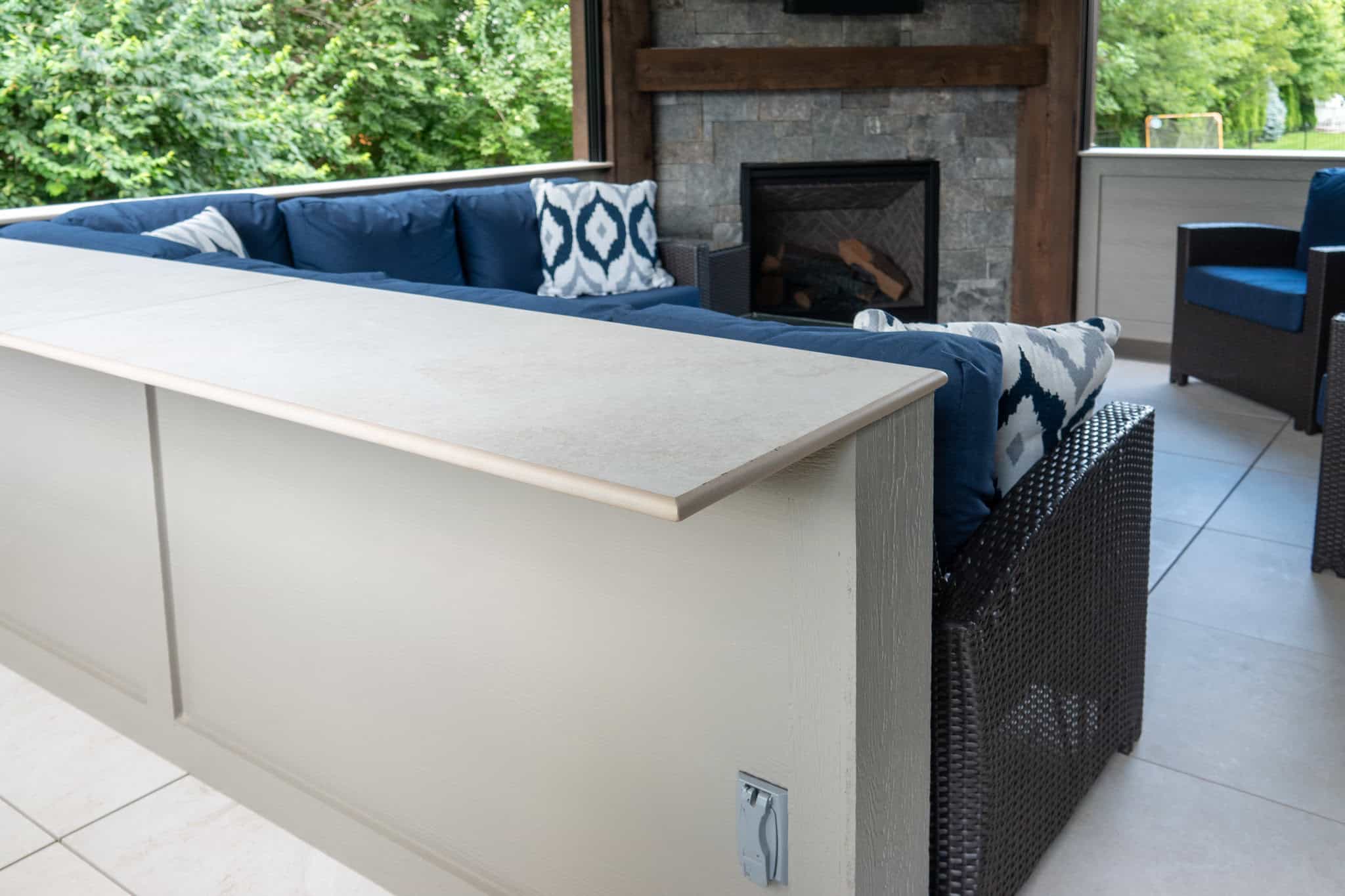 Nicholas Design Build | Outdoor seating area with a stone fireplace, blue cushions, and a tile-topped bar overlooking a green yard.