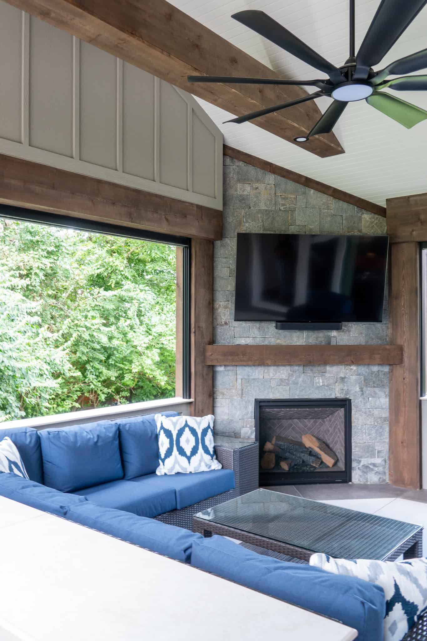 Nicholas Design Build | Outdoor living area with a blue sectional sofa, glass coffee table, wall-mounted TV, and fireplace. Large windows offer a view of greenery outside. Ceiling fan visible above.