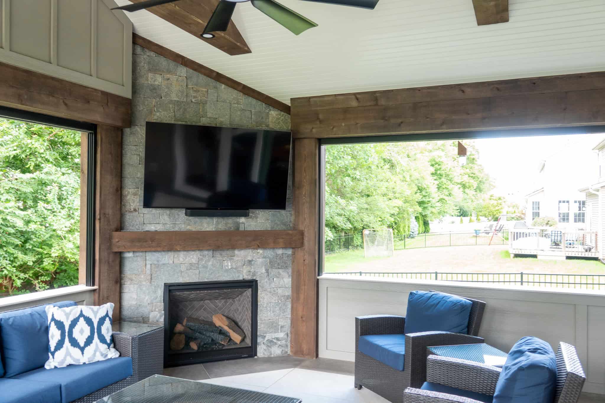 Nicholas Design Build | A cozy outdoor room with a stone fireplace, mounted TV, ceiling fan, and blue cushioned seating. Large windows show a view of green trees and a backyard.