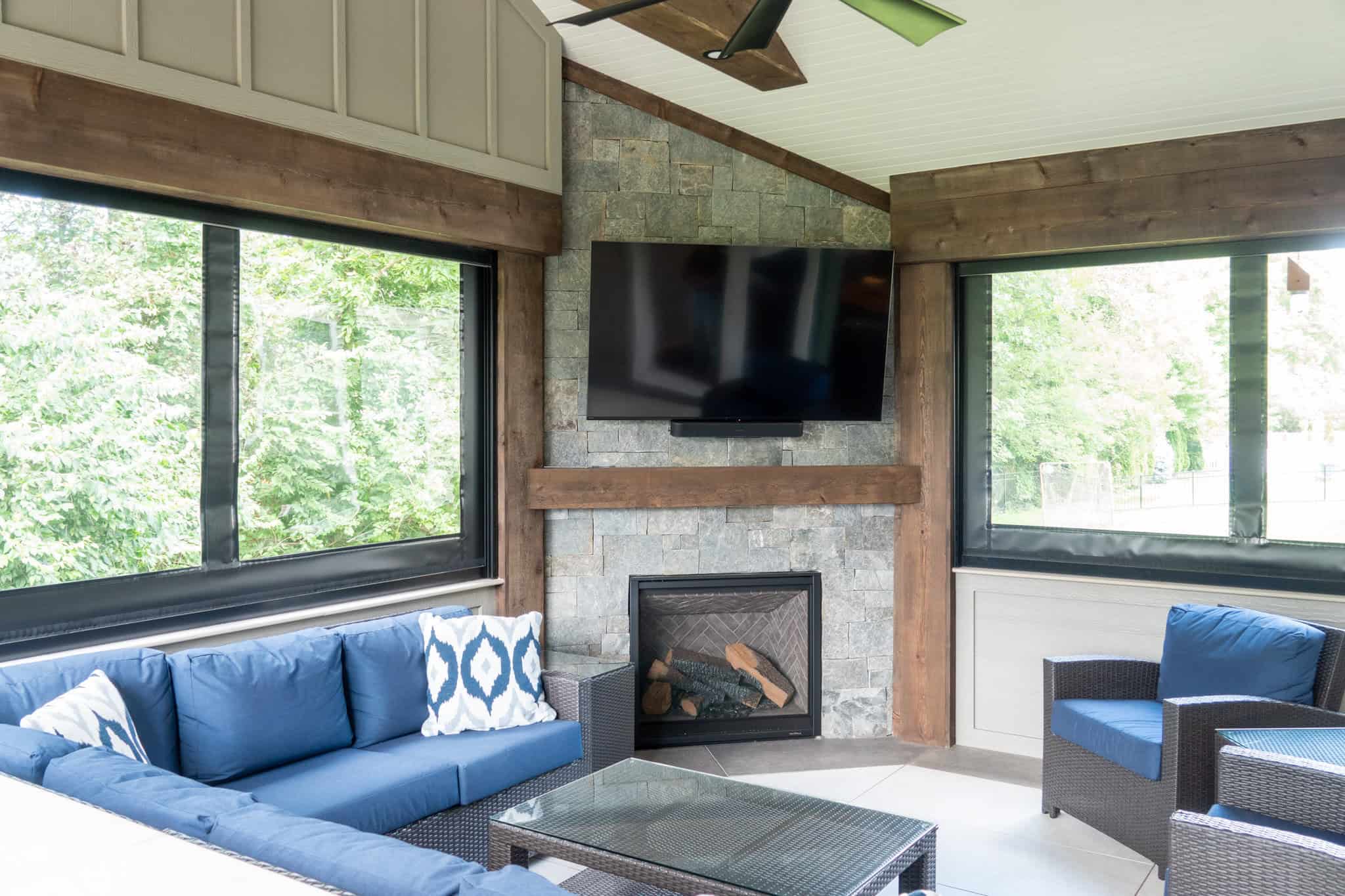 Nicholas Design Build | A cozy indoor patio with a stone fireplace, mounted TV, blue sofas, and large windows offering a view of greenery.