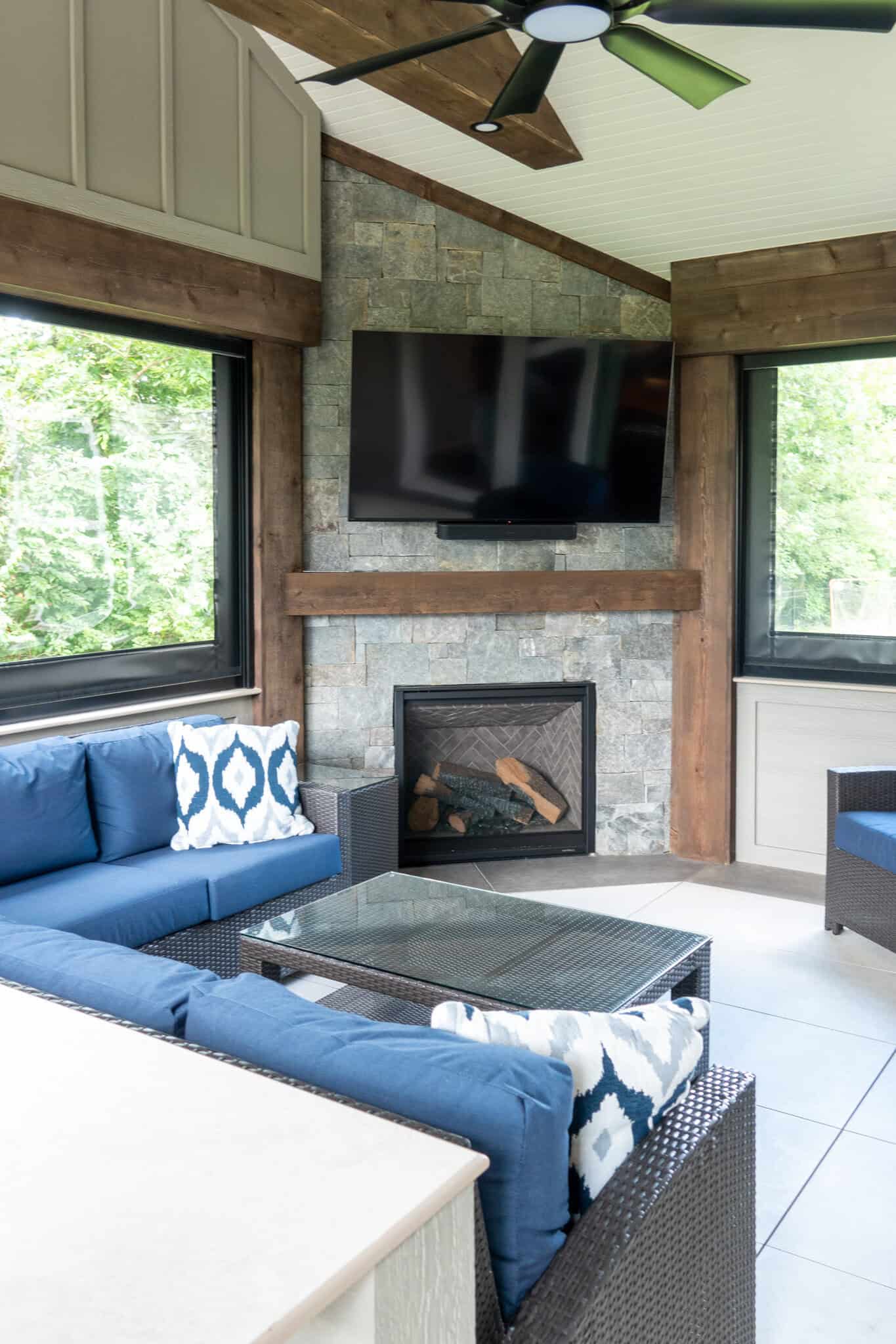 Nicholas Design Build | A sunlit room features a stone wall with wall-mounted TV above a fireplace, surrounded by blue cushioned wicker sofas and a glass-top coffee table. Large windows offer a view of greenery outside.