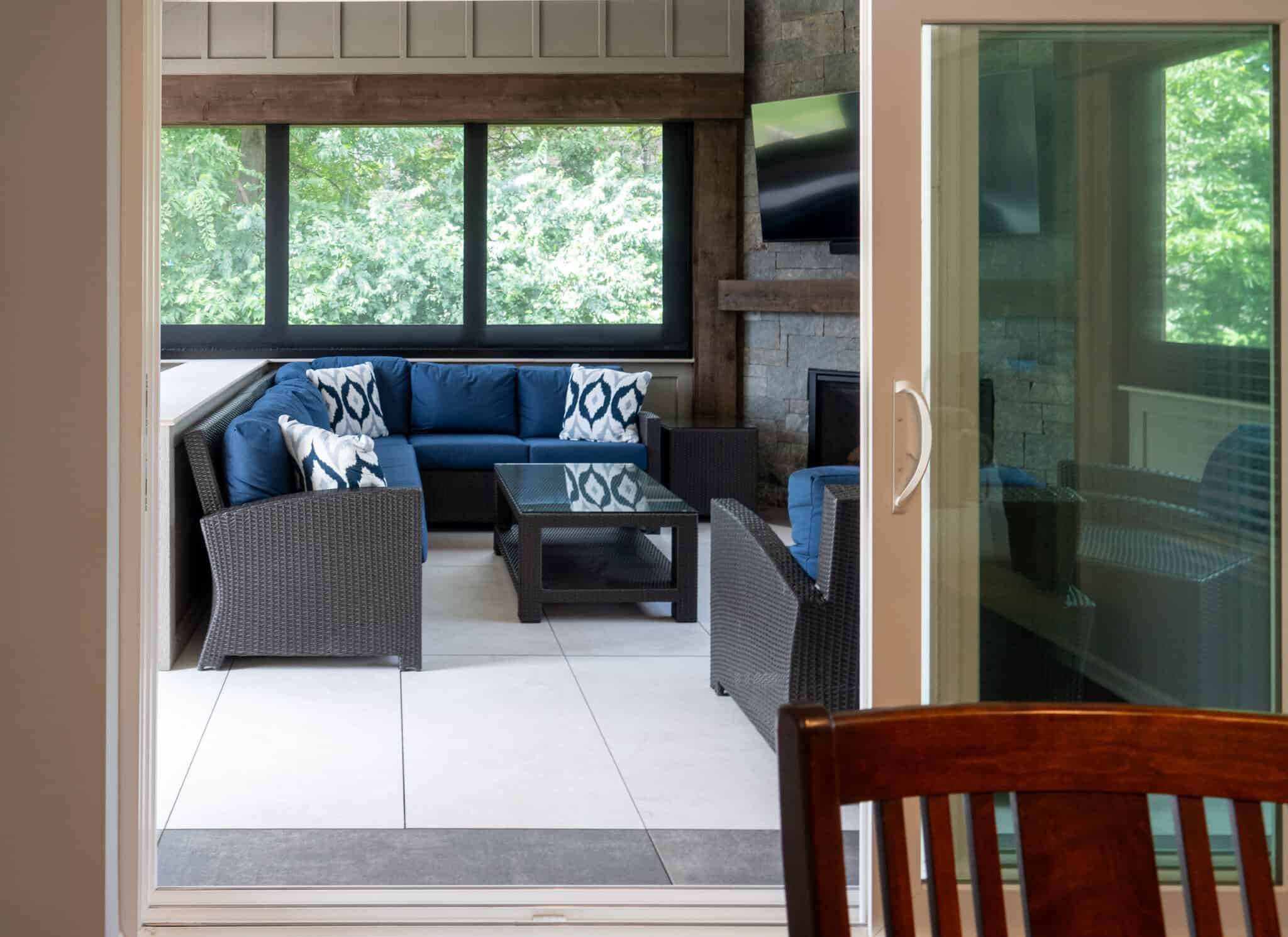 Nicholas Design Build | View of a cozy sunroom with a blue cushioned wicker sofa set, a glass-topped table, and a wall-mounted TV. Large windows offer a view of greenery outside. A wooden chair is partially visible.