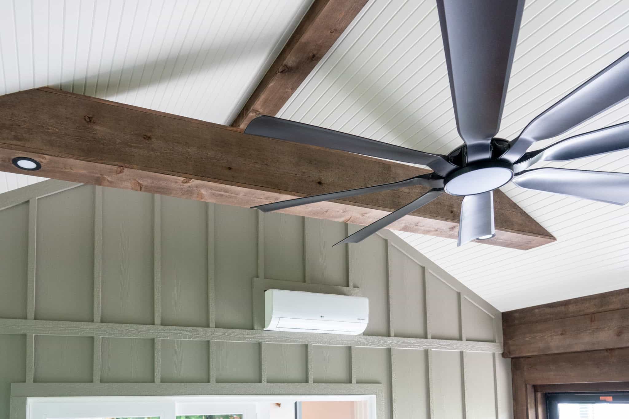 Nicholas Design Build | Ceiling with exposed wooden beams and a modern black ceiling fan, above a beige wall with a white air conditioning unit.