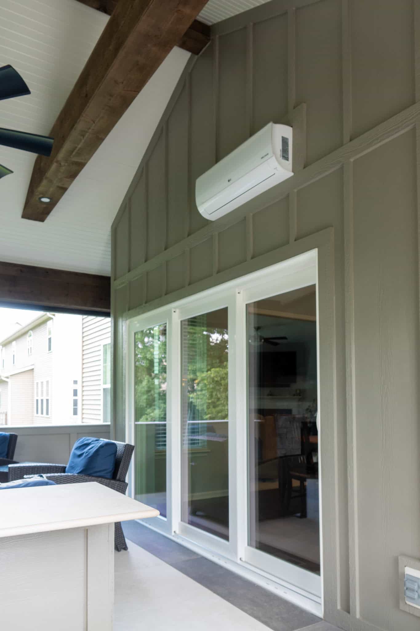 Nicholas Design Build | A porch with wooden beams features a wall-mounted air conditioning unit above sliding glass doors, leading to an interior living area. Chairs and a table are visible outside.