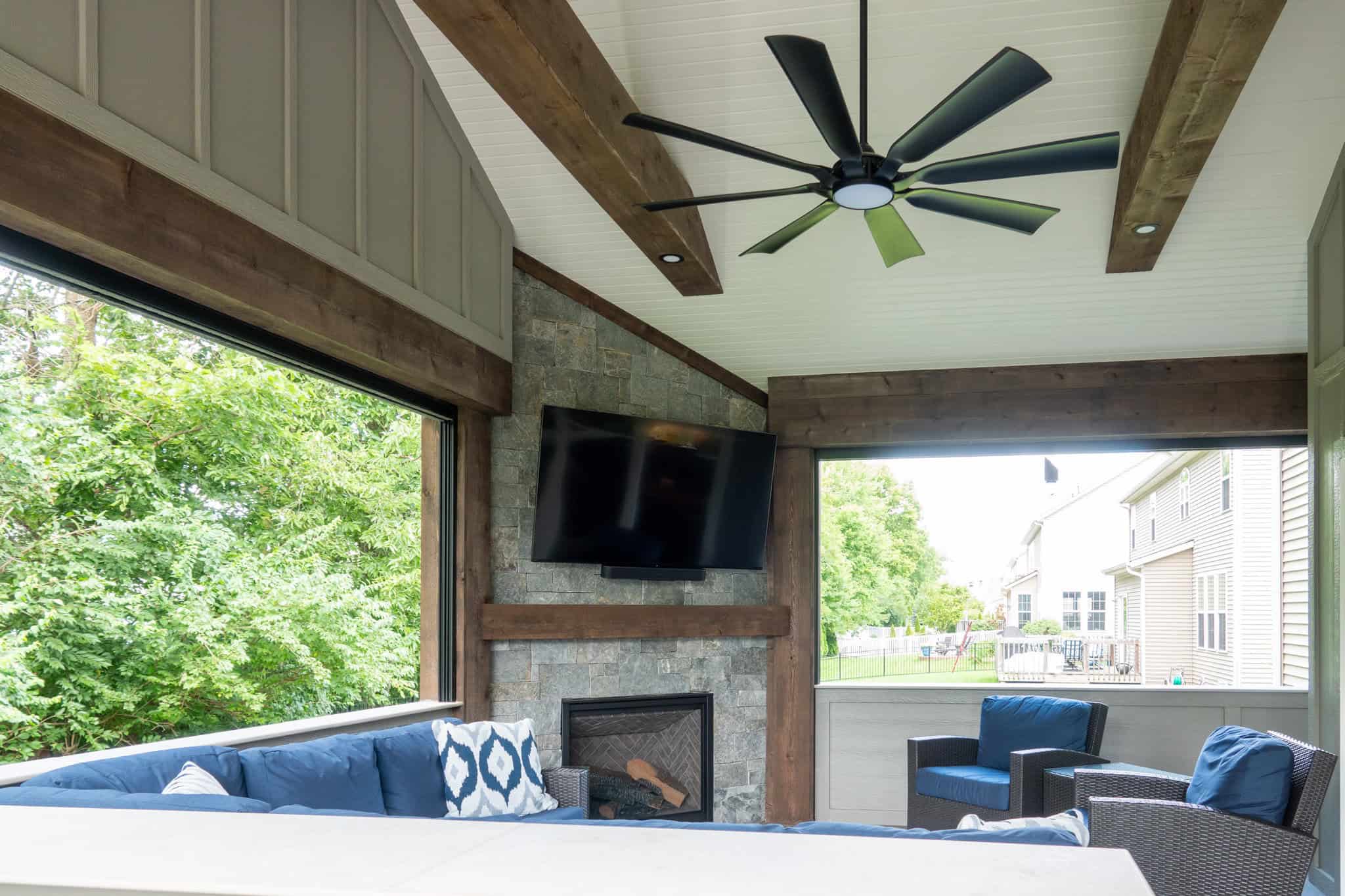 Nicholas Design Build | Covered patio with a ceiling fan, wall-mounted TV, stone fireplace, and blue cushioned seating. Large windows overlook a green yard and neighboring houses.