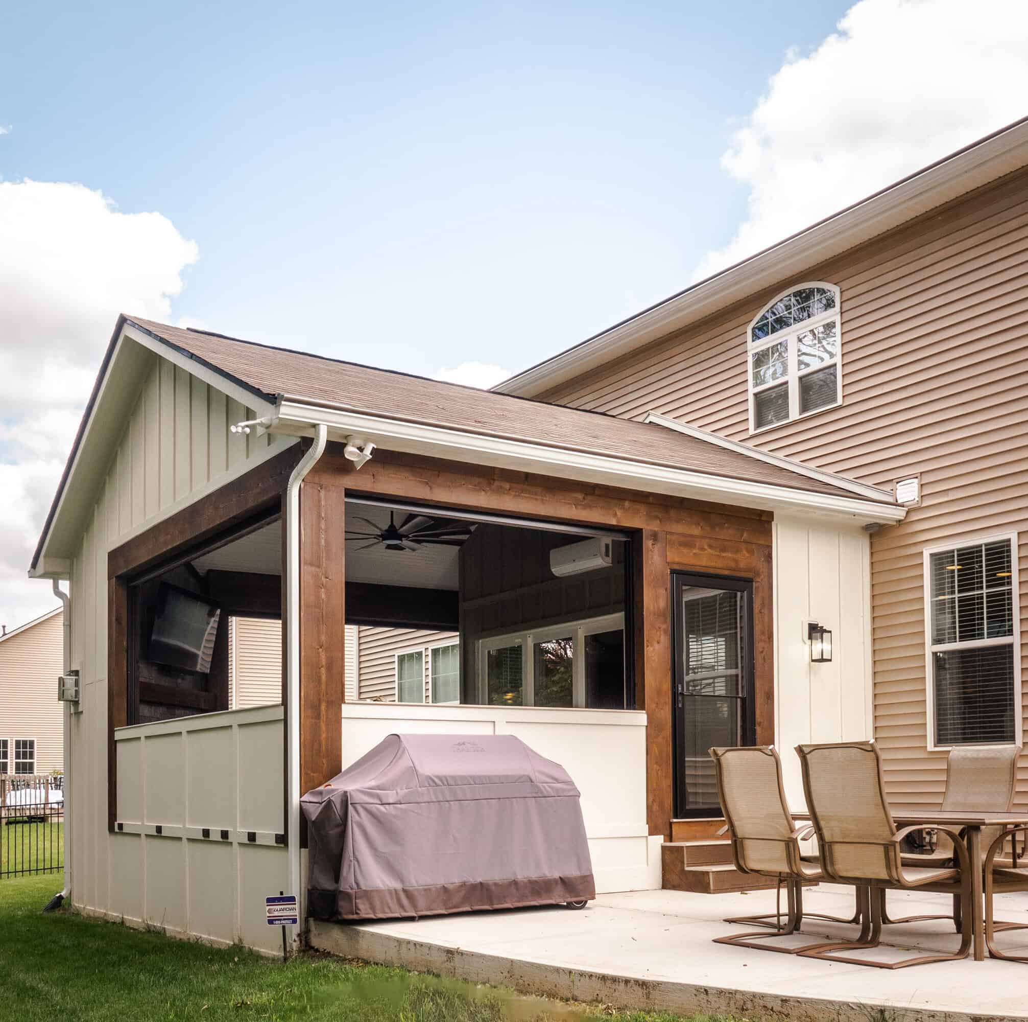 Nicholas Design Build | A covered patio with a grill and outdoor dining set attached to a beige house. The area is enclosed with white and wood paneling.