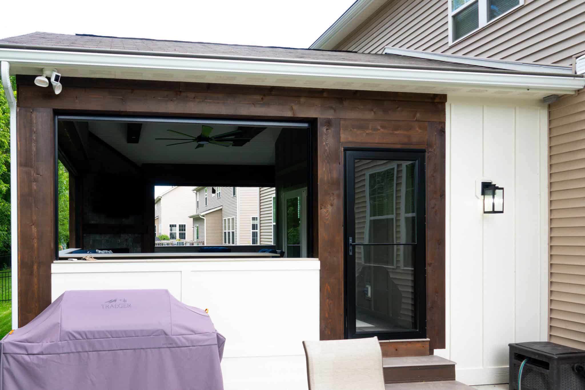 Nicholas Design Build | Outdoor patio with a covered barbecue area, a grill, a ceiling fan, and seating. A window and door are on the side of a house with tan siding and white trim.