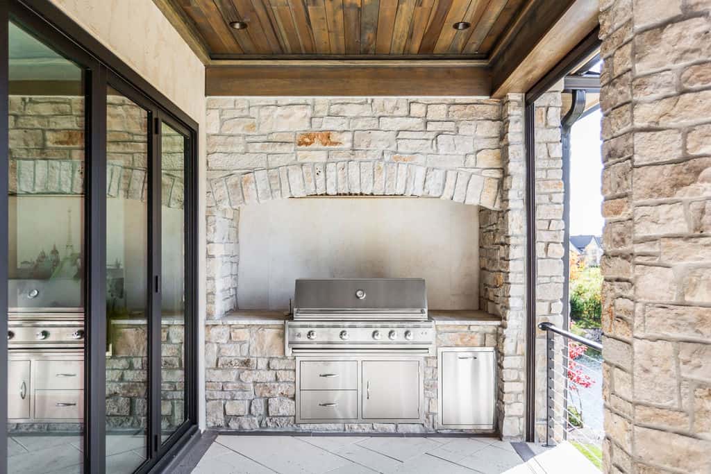 Nicholas Design Build | Outdoor stone kitchen with a built-in stainless steel grill and storage drawers. Large sliding glass doors and wooden ceiling are visible.