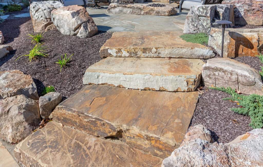 Nicholas Design Build | Stone steps in a garden setting, surrounded by large rocks, mulch, and small plants.