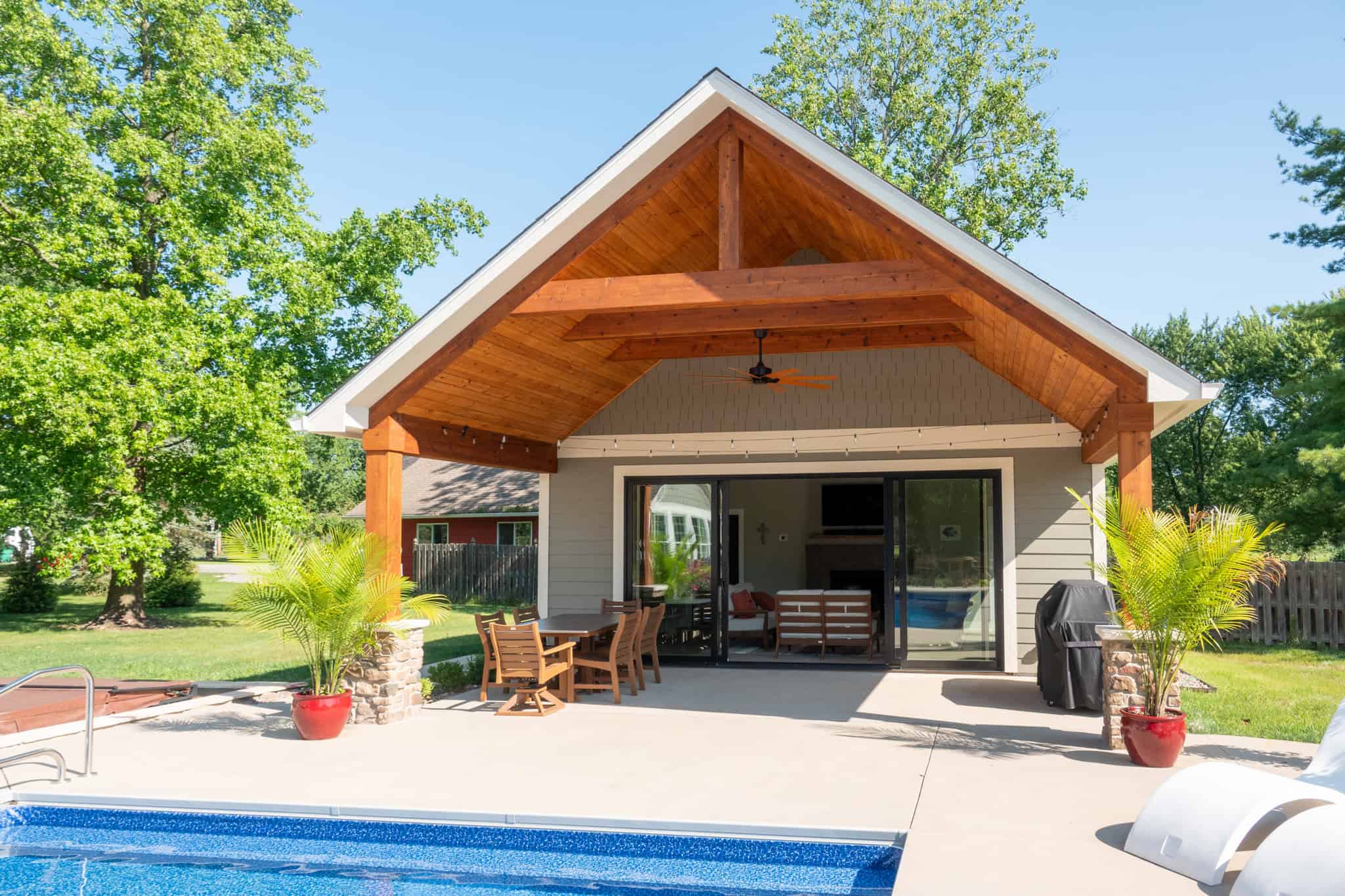 Nicholas Design Build | A backyard features a patio with wooden furniture, a shaded structure, and an adjacent pool. Trees and plants surround the area.