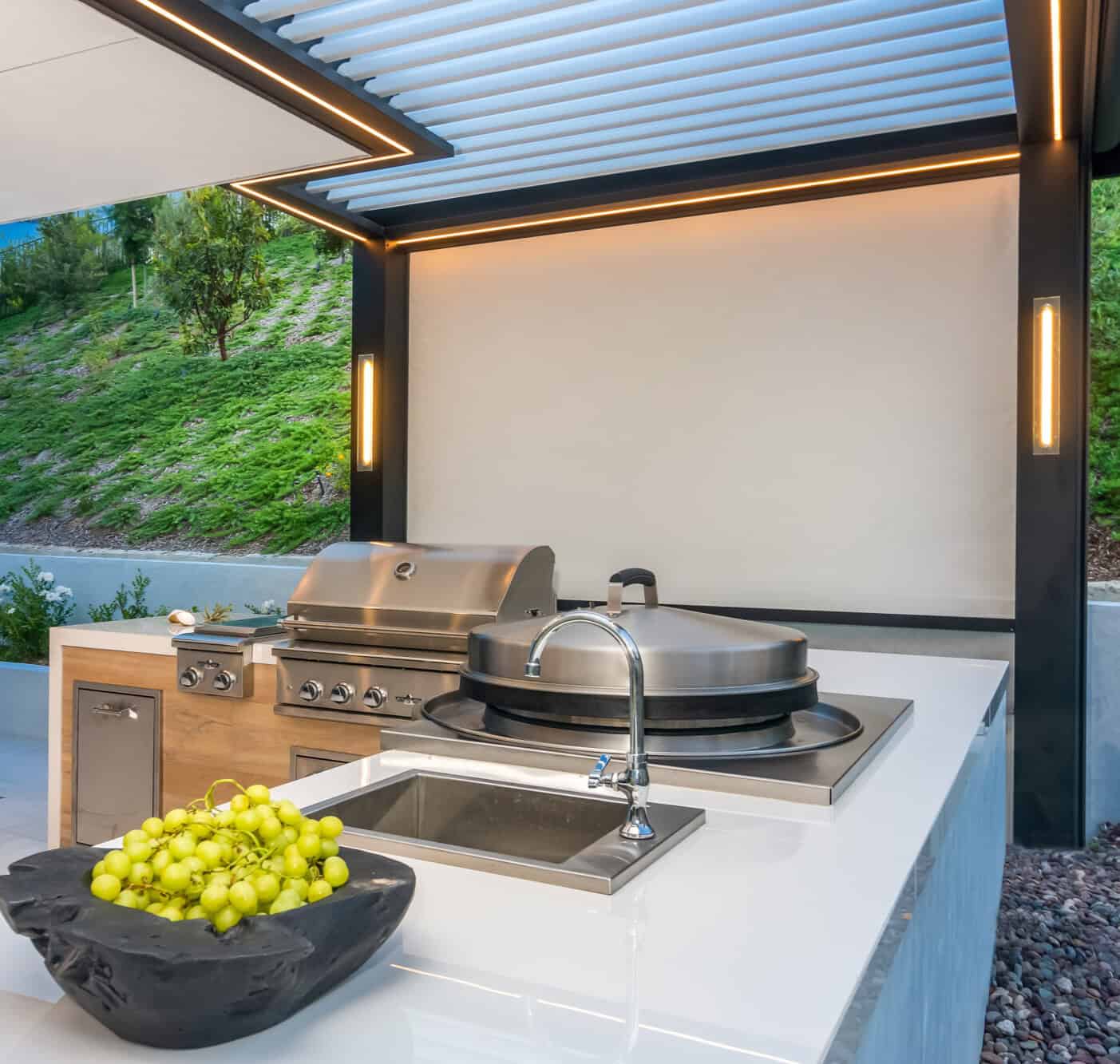 Nicholas Design Build | Outdoor kitchen with stainless steel grill and rotisserie, sink, and a bowl of green grapes on a white countertop. Overhead lighting and modern design elements are visible.