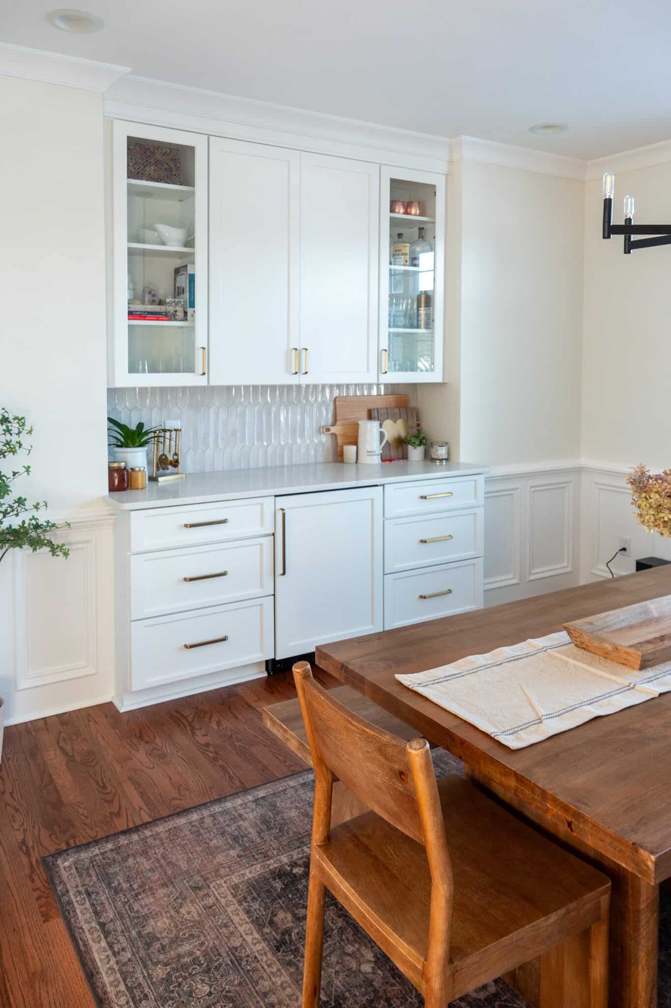 Nicholas Design Build | Dining area with a wooden table, a chair, and a sideboard. The sideboard has white cabinets with glass fronts, decorated with plants and kitchen items. A patterned rug sits on a wooden floor.