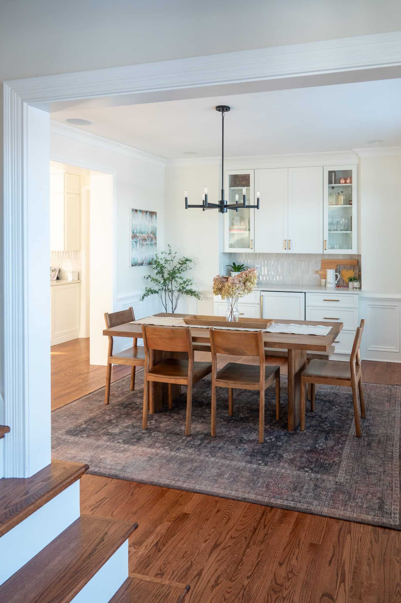 Nicholas Design Build | A dining room with a wooden table and six chairs on a rug, beneath a modern chandelier. A sideboard with cabinets is in the background, with decor including a potted plant and framed art.