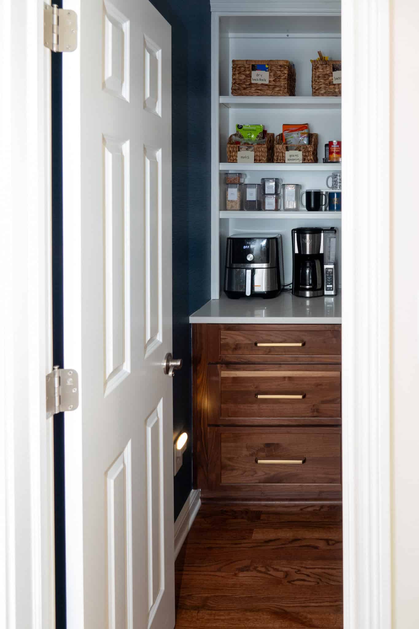 Nicholas Design Build | Open pantry door reveals wooden shelves with wicker baskets, spice jars, an air fryer, coffee maker, and various kitchen items. Dark cabinets and wooden floor complete the scene.