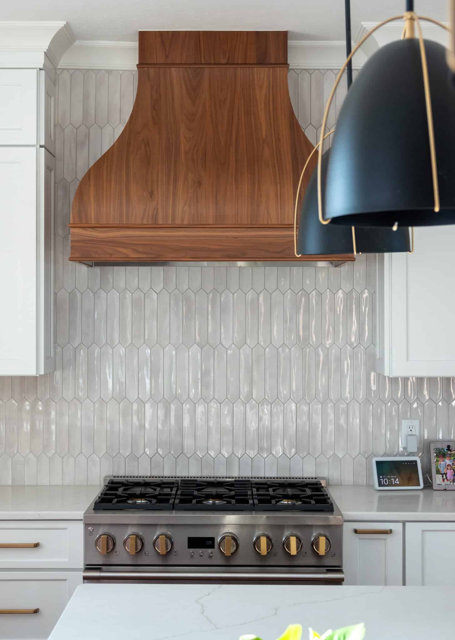 Nicholas Design Build | Modern kitchen with a stainless steel stove, wooden range hood, and geometric tile backsplash. Black pendant light in the foreground.