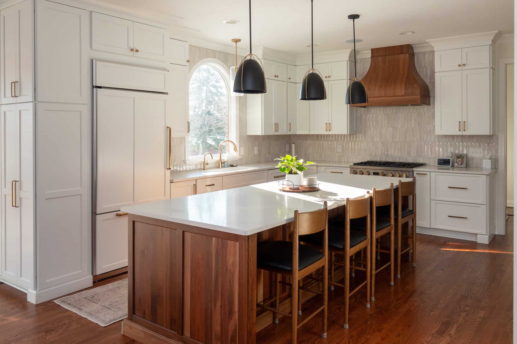 Nicholas Design Build | Modern kitchen with white cabinets, a wooden island with barstools, pendant lights, and a wooden range hood. Large arched window provides natural light. Dark wood floor adds contrast.