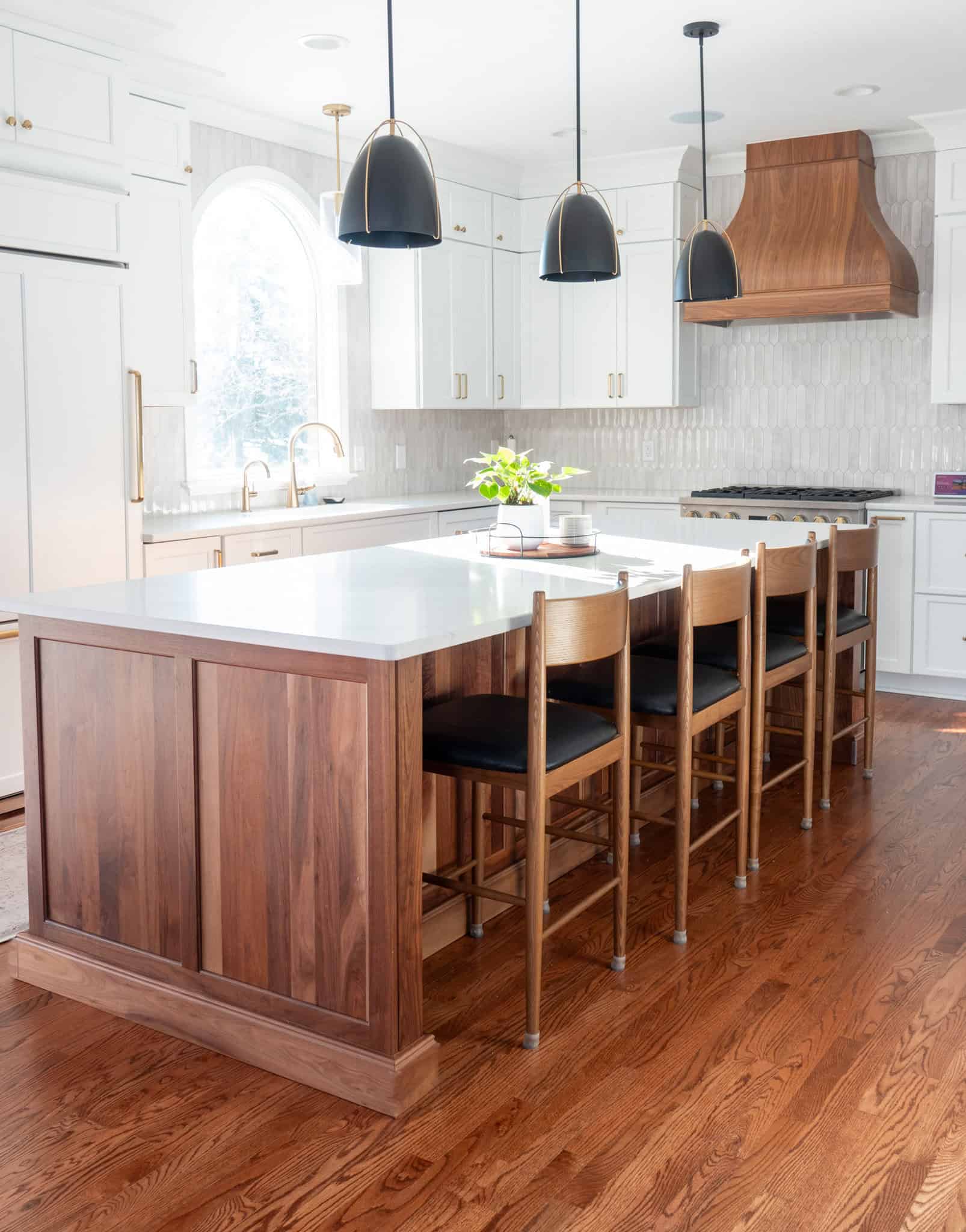 Nicholas Design Build | A modern kitchen with a wooden island, four chairs, white cabinetry, black pendant lights, hardwood floor, and a window.