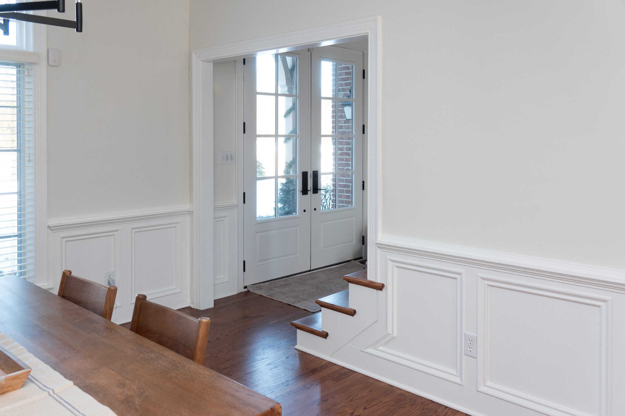 Nicholas Design Build | A dining area with a wooden table and chairs is set beside a doorway with glass-paneled double doors, leading outside. The room has white wainscoting and hardwood floors.