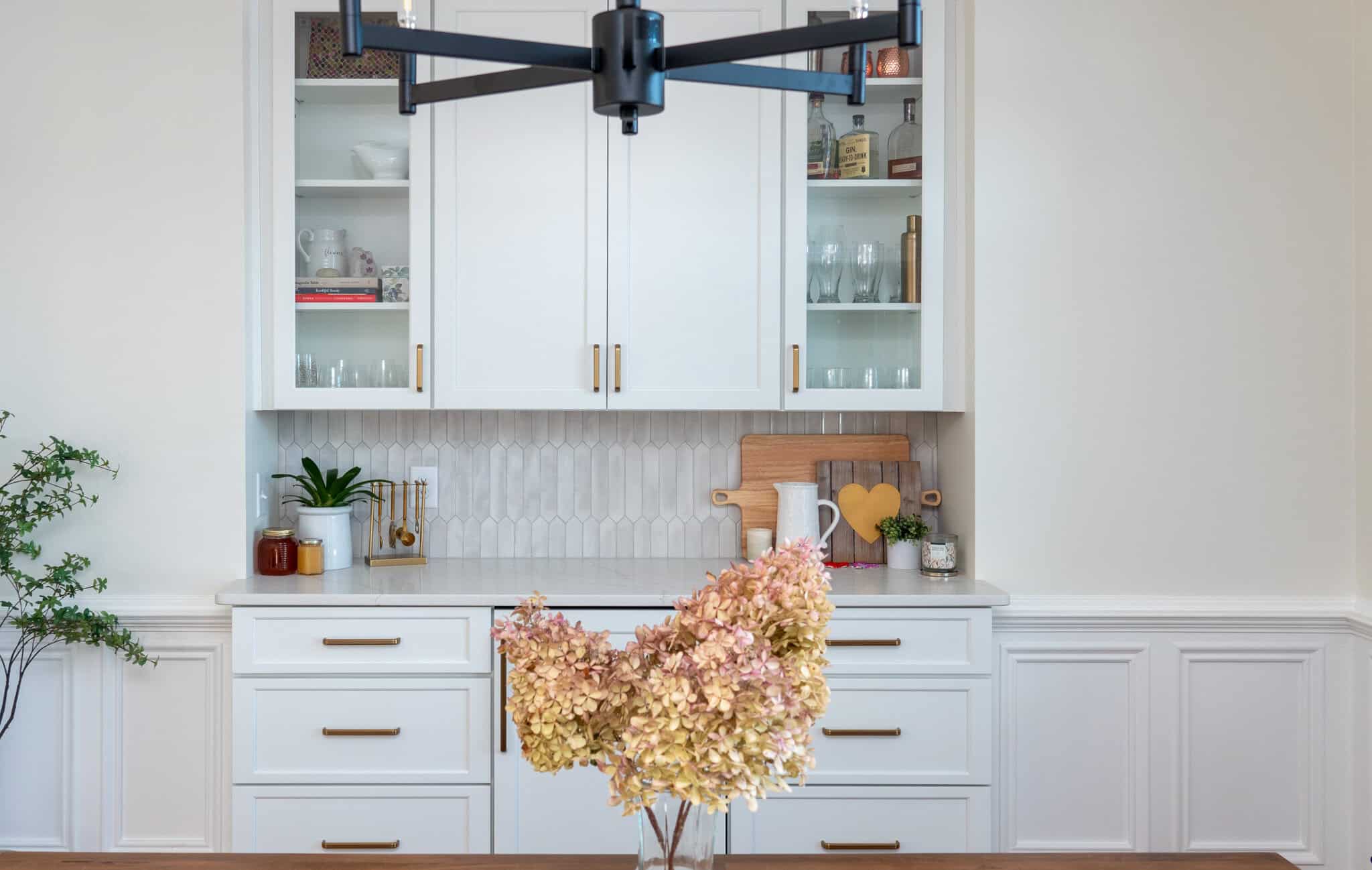 Nicholas Design Build | A kitchen area with white cabinets and drawers, glass cupboard doors, decorative plants, and a vase with dried hydrangeas on the counter. A modern black chandelier hangs above.