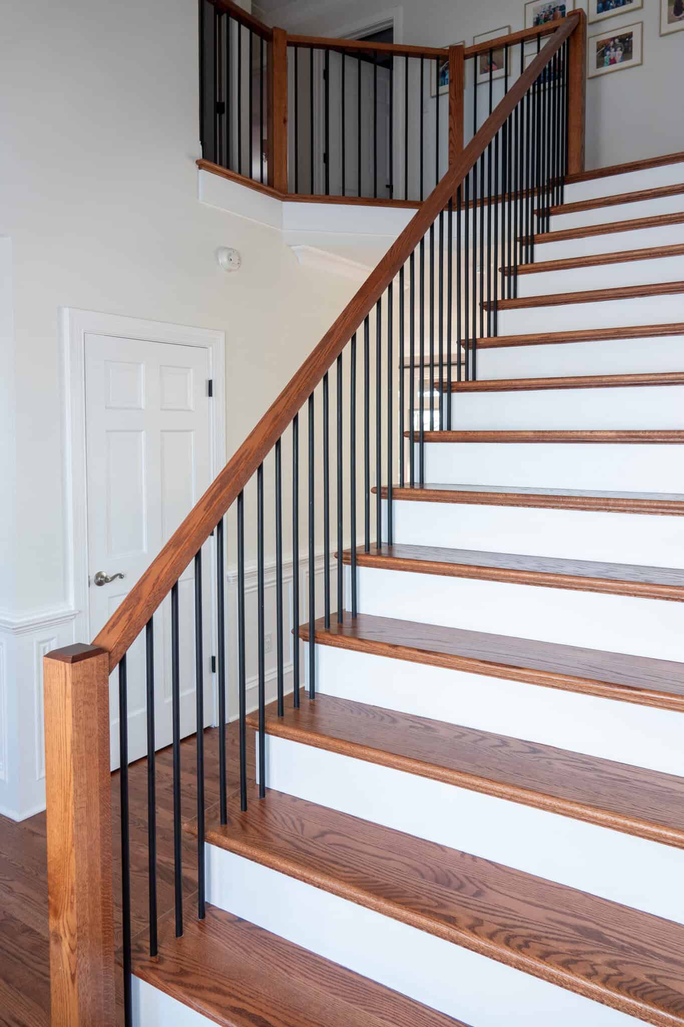 Nicholas Design Build | Wooden staircase with black metal balusters leading to the upper floor of a house, white walls, and several framed photos on the wall.