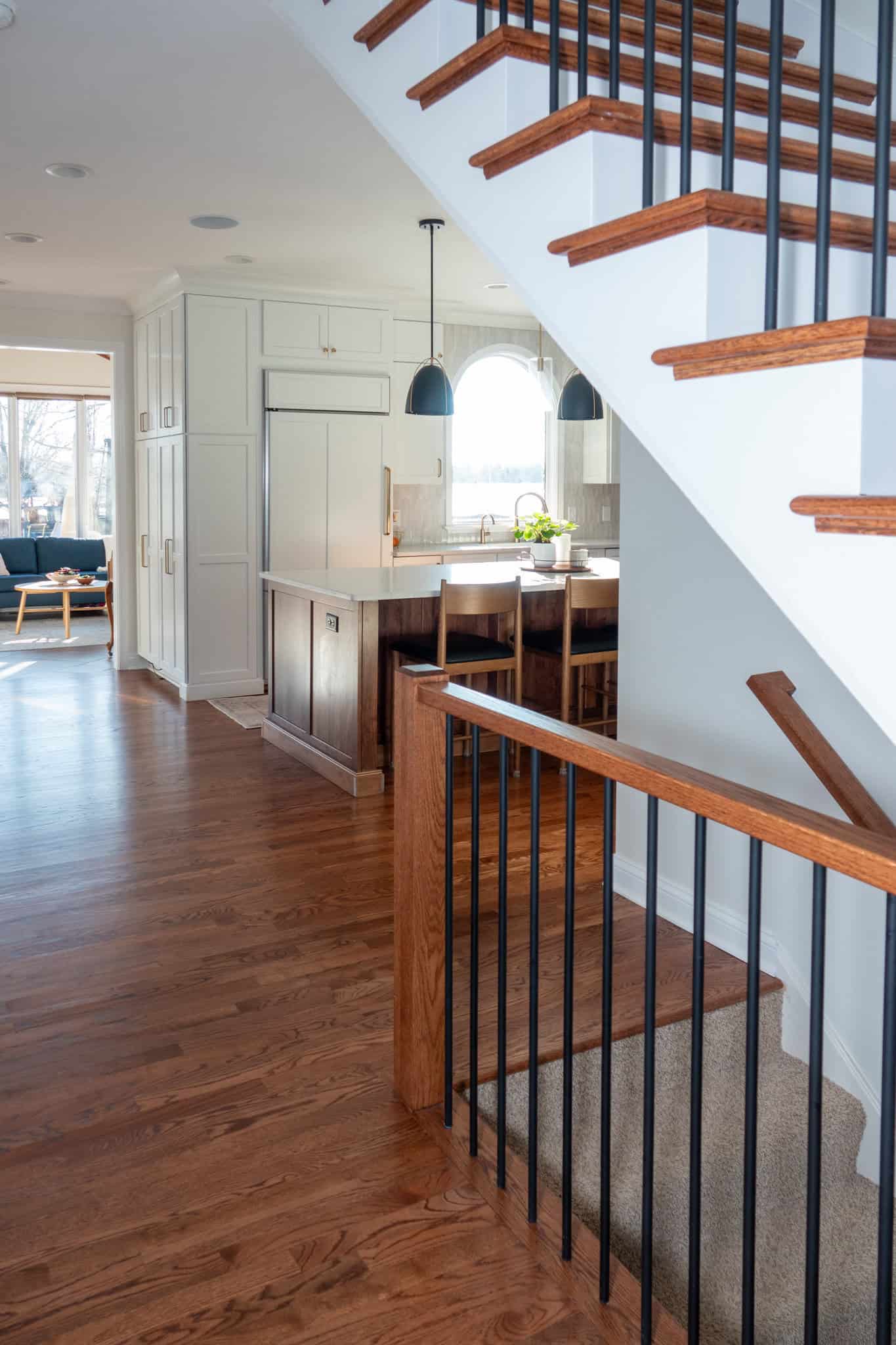 Nicholas Design Build | A modern kitchen with an island, pendant lights, and wooden cabinetry is visible from a staircase with wooden steps and black railings. A living area is seen through a doorway.