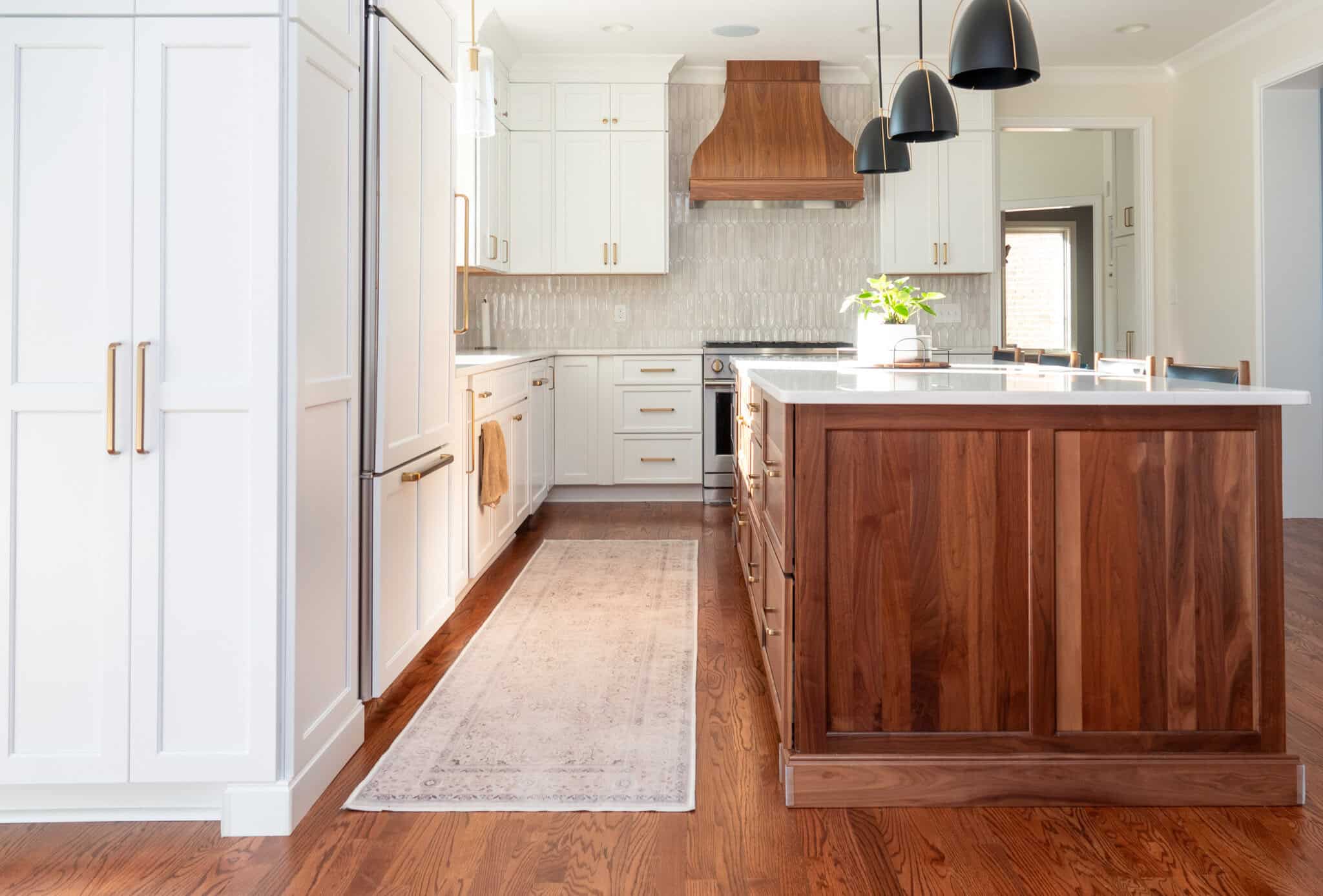 Nicholas Design Build | Modern kitchen with white cabinets, wood island, and stainless steel appliances. Pendant lights hang above the island, and a rug runs along the wooden floor.