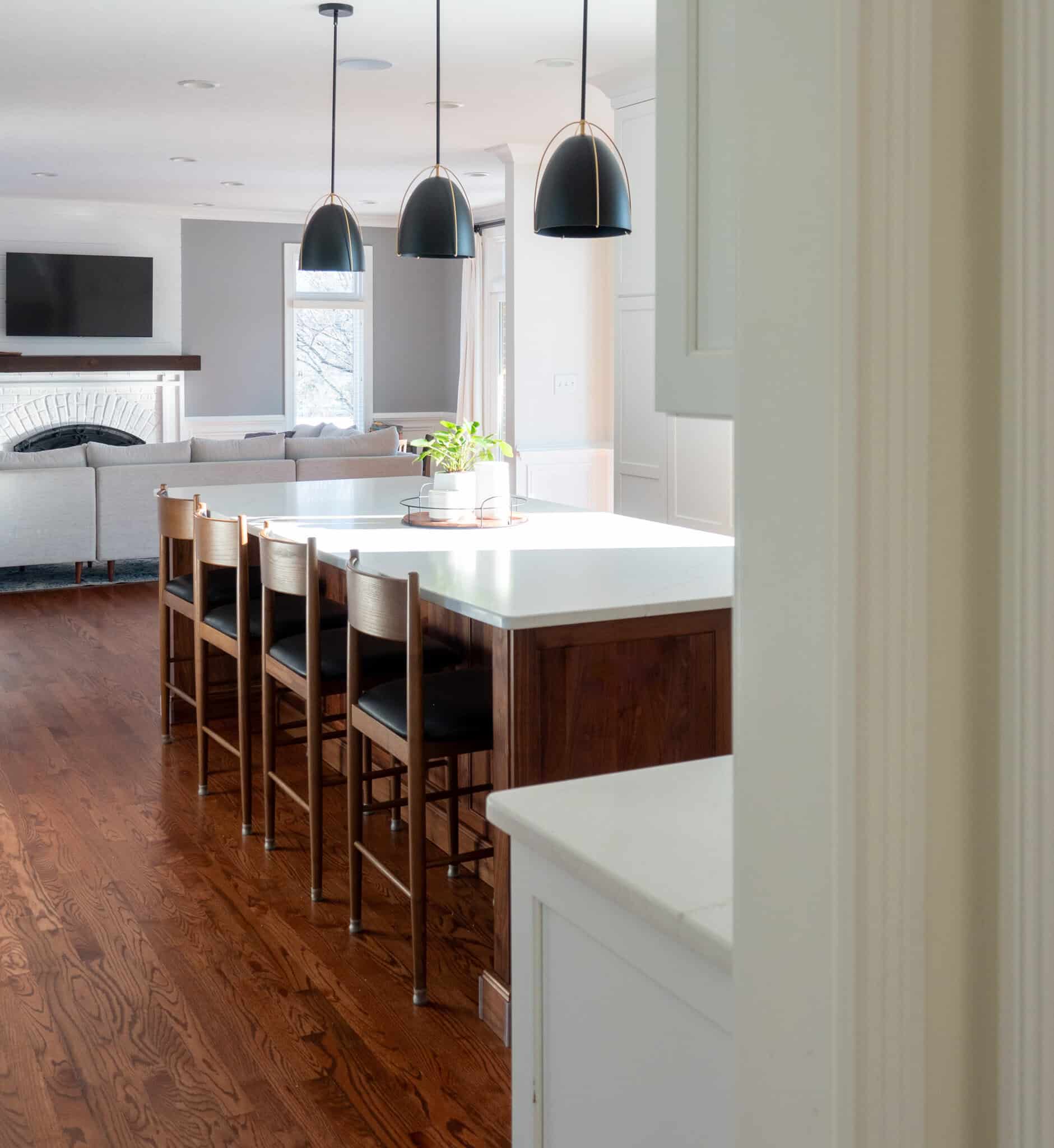 Nicholas Design Build | Modern kitchen with a wooden island and white countertop, surrounded by four chairs. Three pendant lights hang above. A living room with a TV and fireplace is in the background.