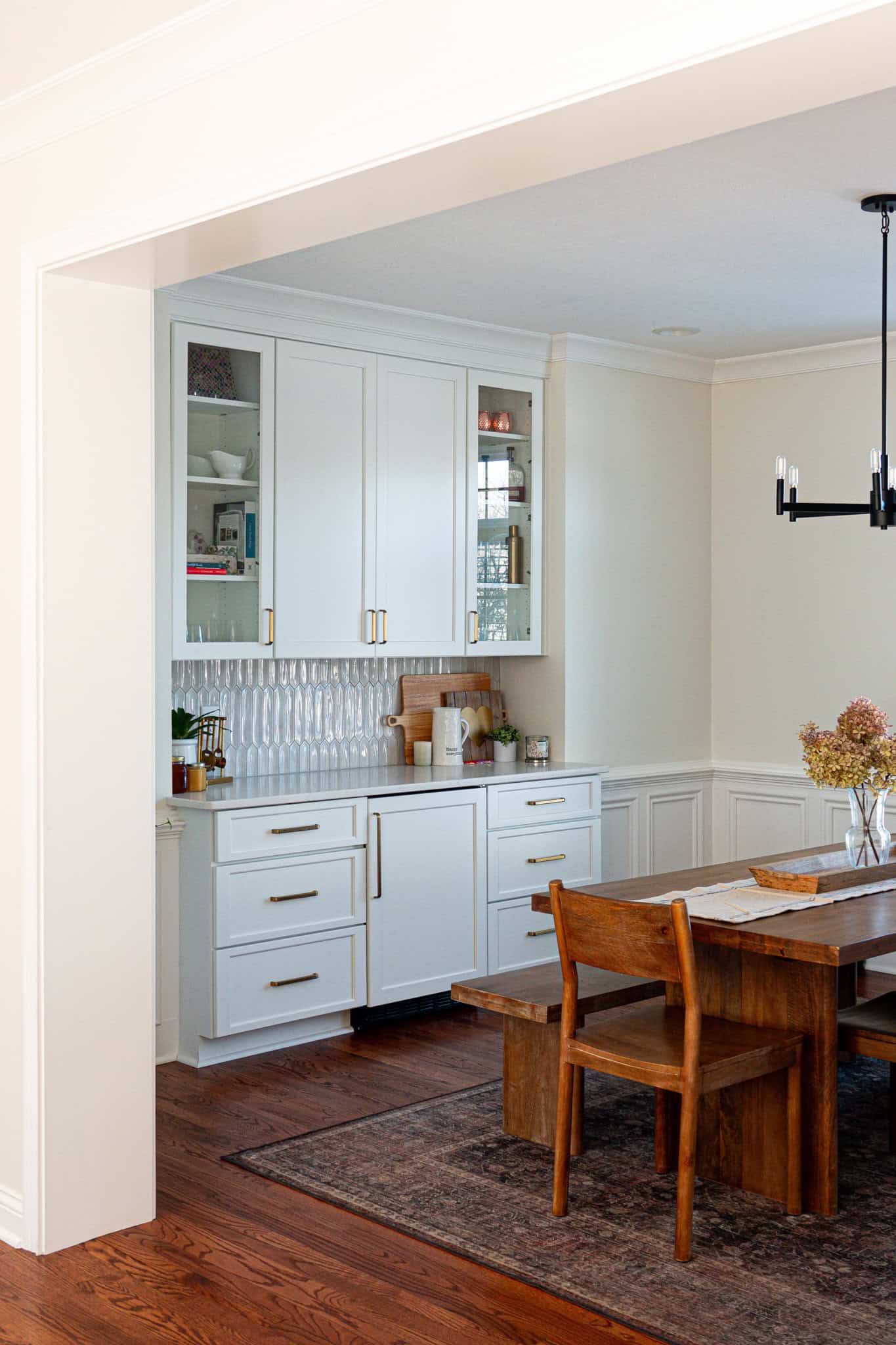 Nicholas Design Build | A dining area with a wooden table and chairs, a modern chandelier, and a white cabinet with a textured backsplash. A rug is under the table on hardwood flooring.