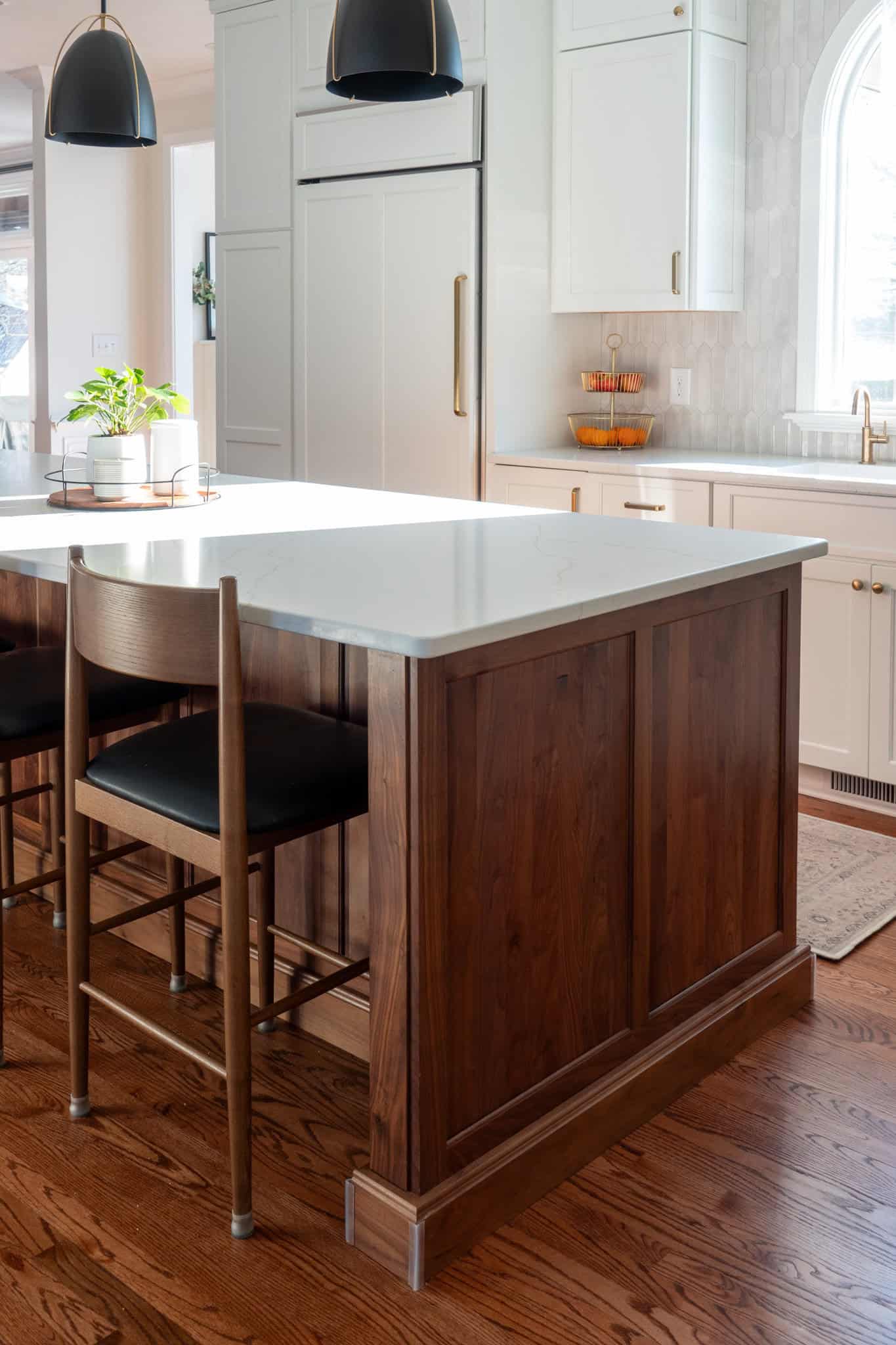 Nicholas Design Build | Modern kitchen with a wooden island, black barstools, white cabinets, pendant lights, and a window. A plant and fruit stand are on the counter. Light wood flooring.