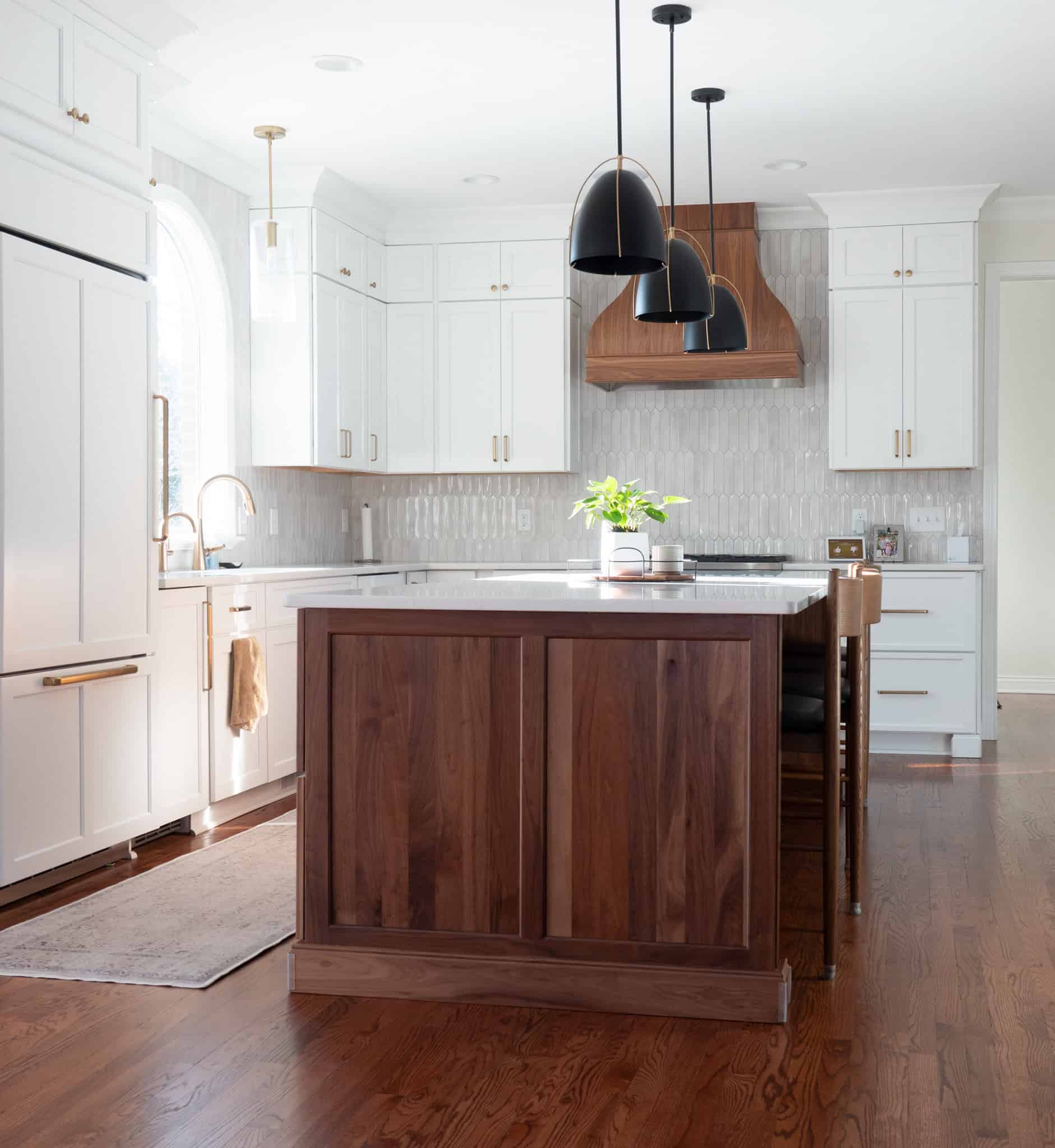 Nicholas Design Build | Modern kitchen with white cabinets, a wooden island, and dark pendant lights. Wood floors and marble backsplash accentuate the space. A potted plant sits on the island countertop.