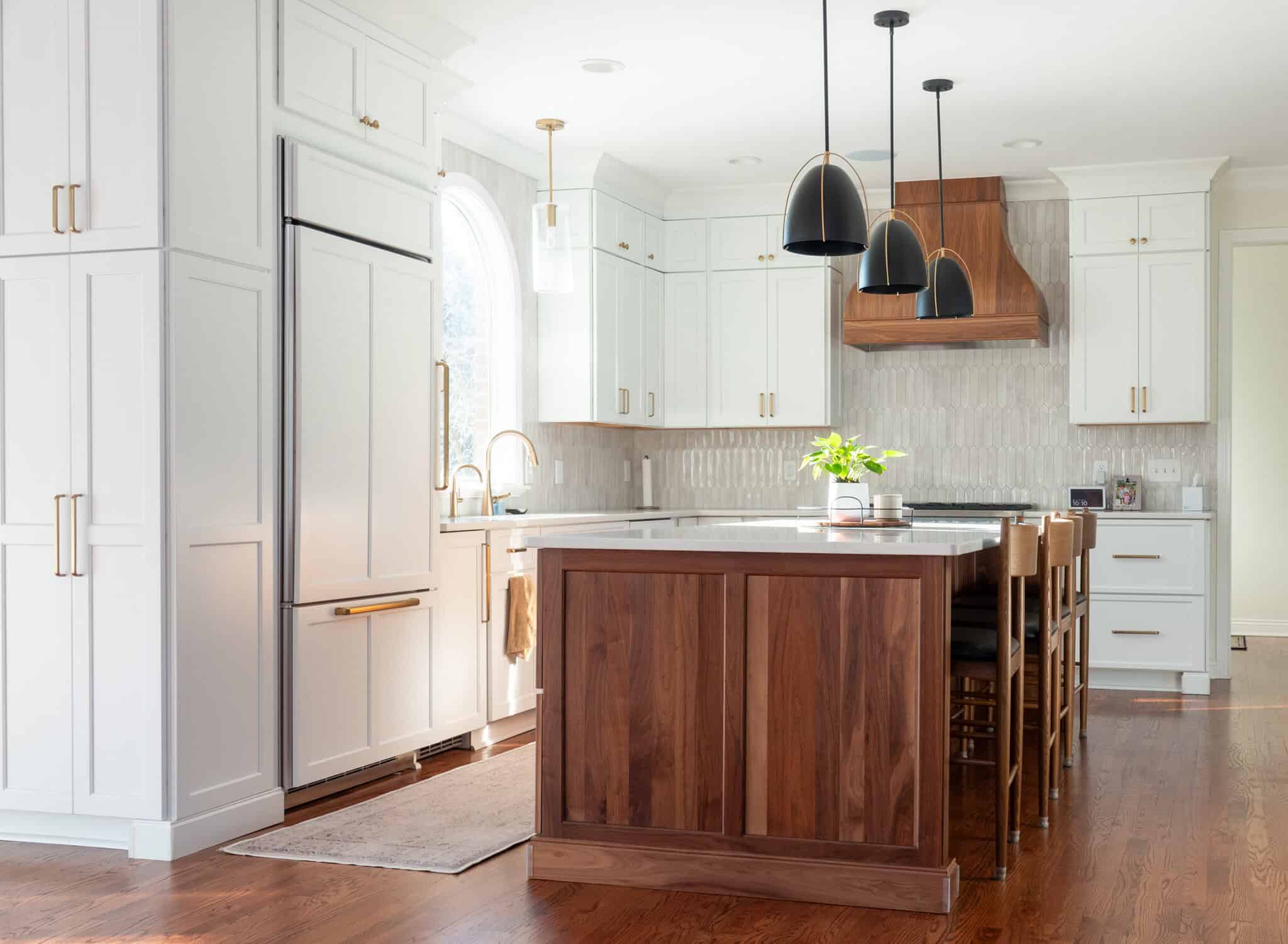 Nicholas Design Build | Modern kitchen with white cabinets, a wood island, and black pendant lights. Stainless steel appliances and wooden bar stools are present. Large window above the sink allows natural light.