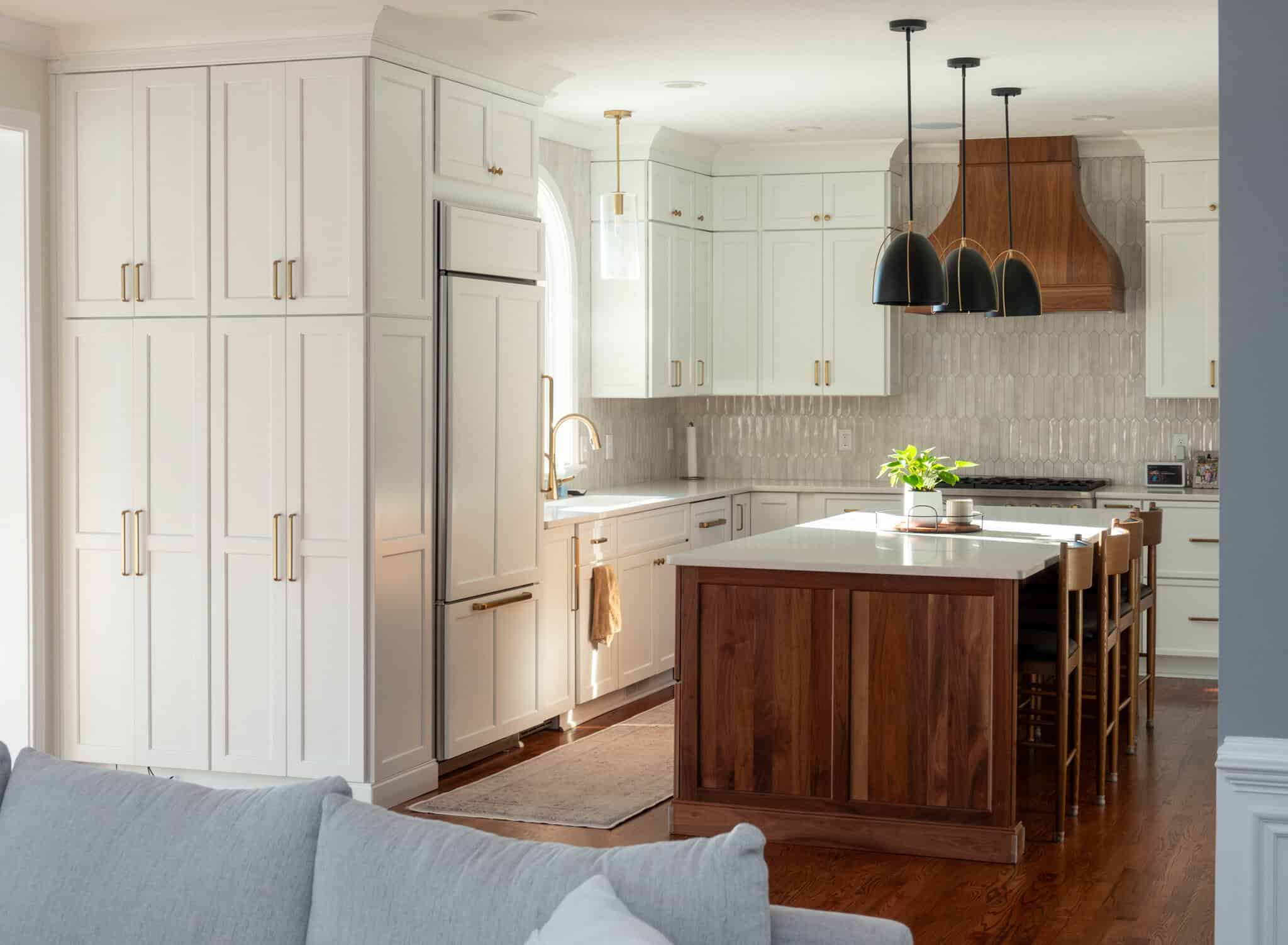 Nicholas Design Build | A modern kitchen with white cabinets, a center island, and wooden accents. Several pendant lights hang above the island. A plant decorates the countertop.