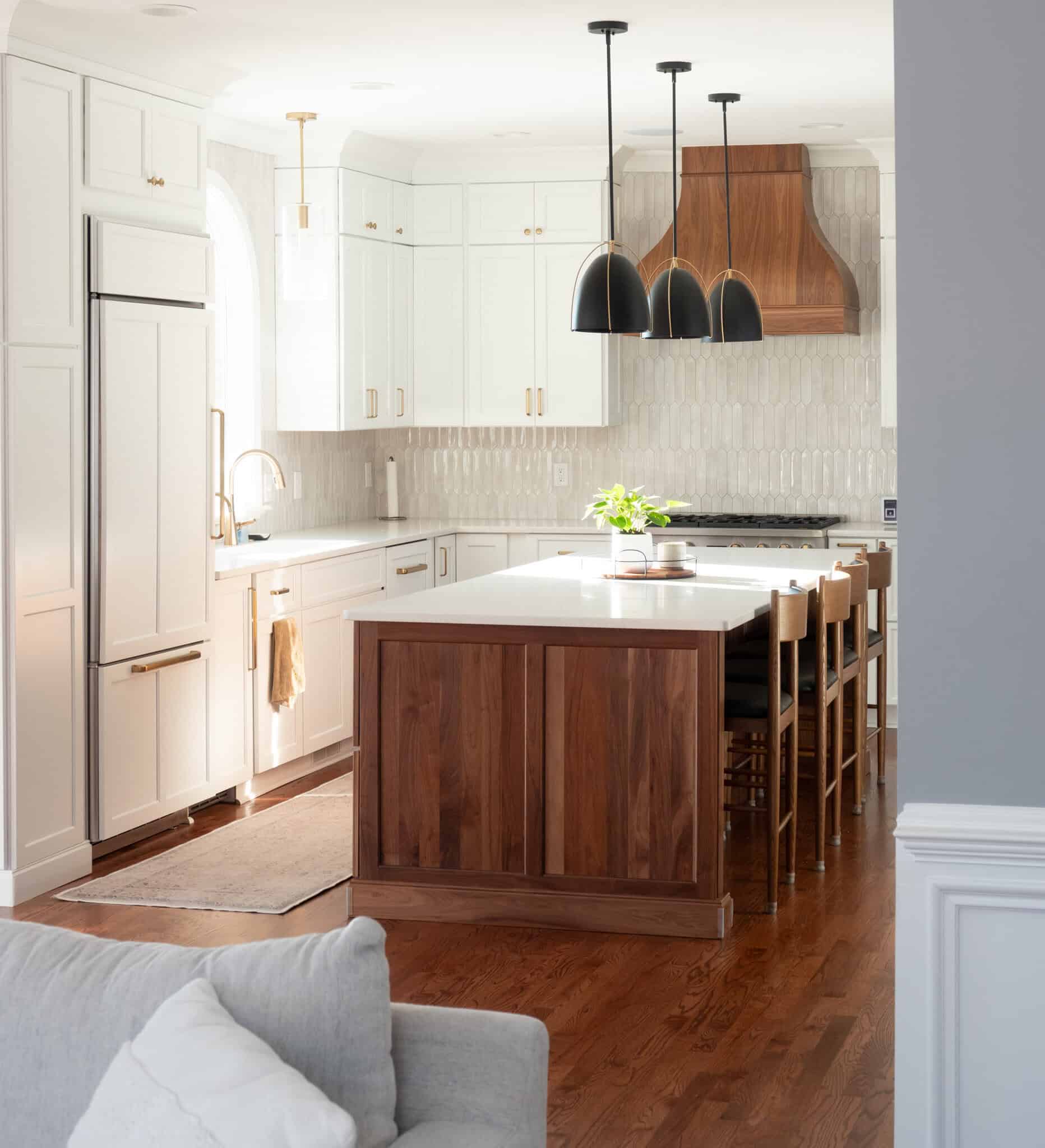 Nicholas Design Build | Modern kitchen with white cabinetry, wooden island with stools, pendant lights, and a wooden range hood. Light enters through a large window, illuminating the hardwood floor.