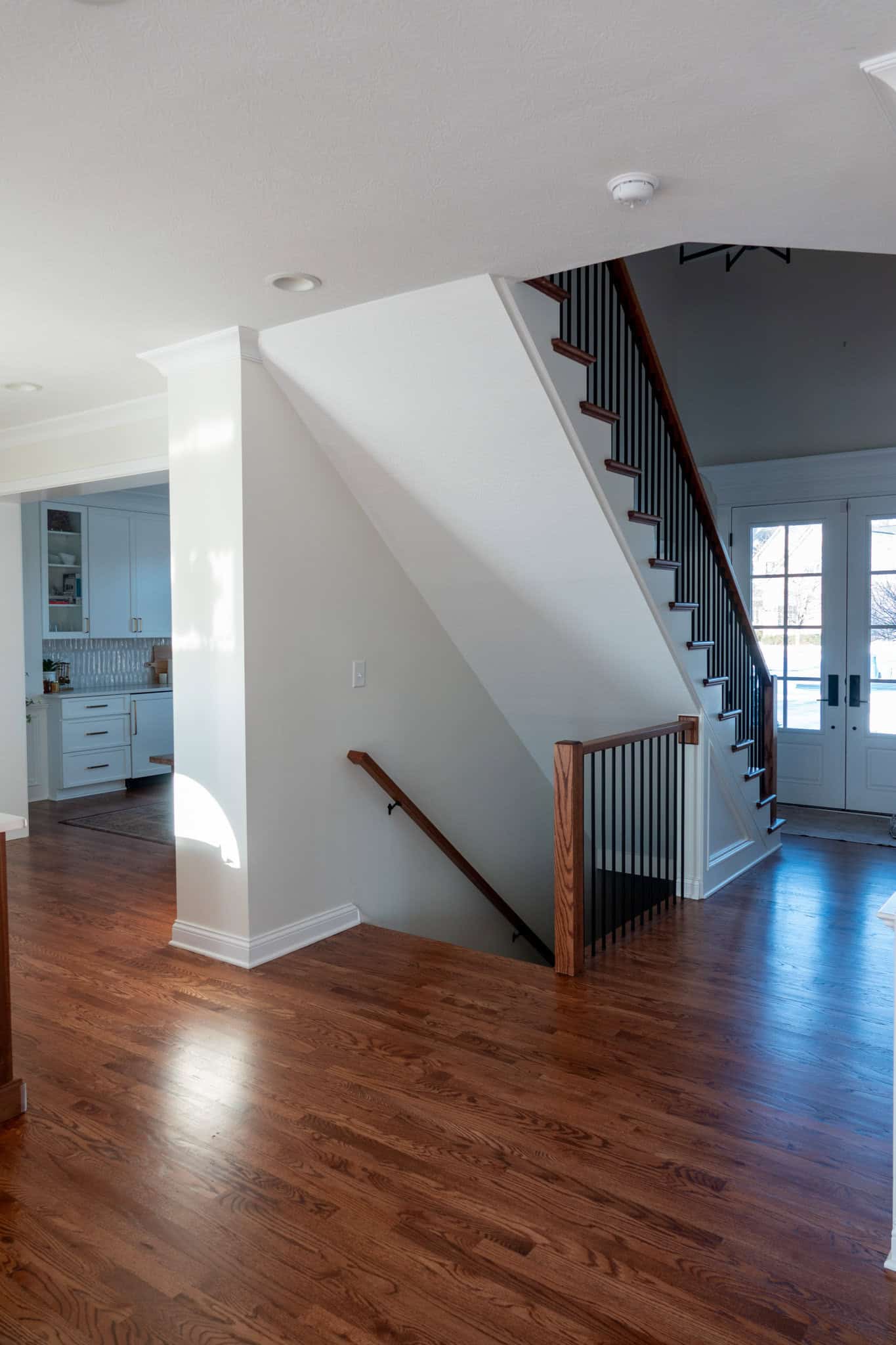 Nicholas Design Build | A wooden staircase with black railings descends in a bright hallway with hardwood floors, leading to a kitchen and double doors with glass panels.